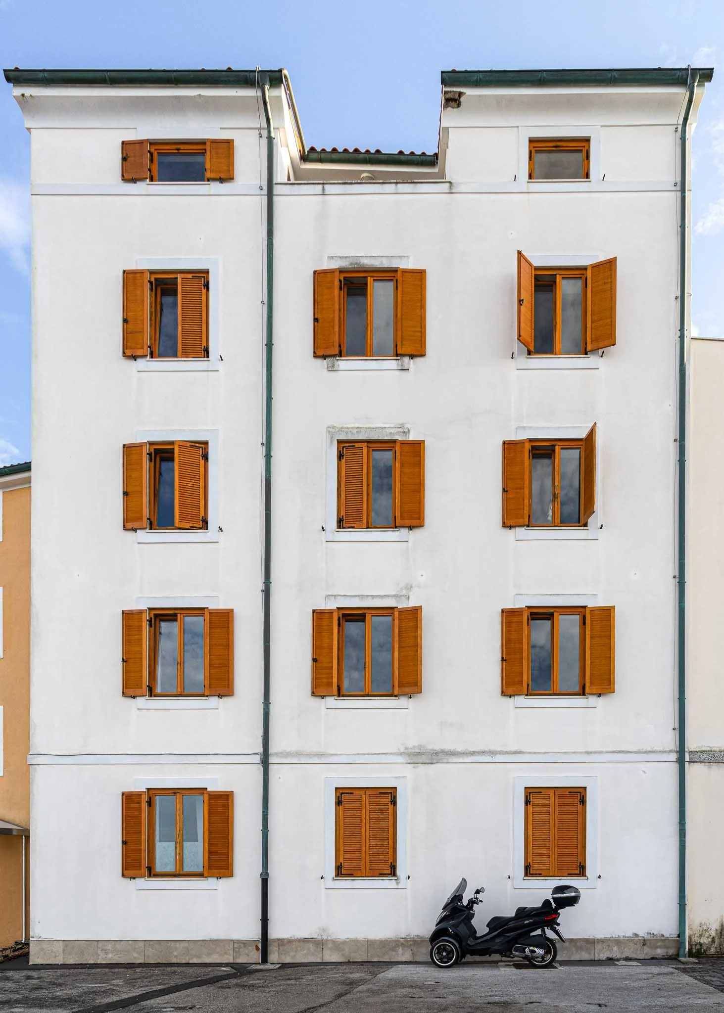 Symmetrical white building facade with rows of orange wooden window shutters and a black scooter parked below, photographed in Slovenia.
