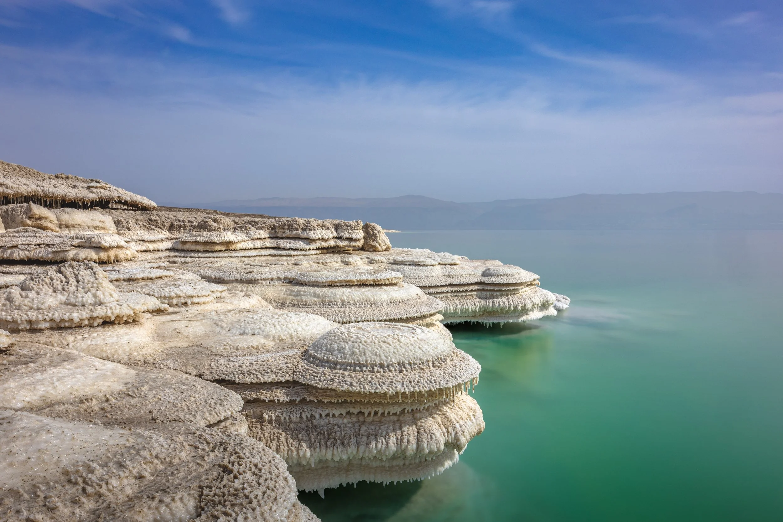 Salt formations along the edge of a tranquil, turquoise lake with a mountainous landscape in the background and a partly cloudy sky overhead.