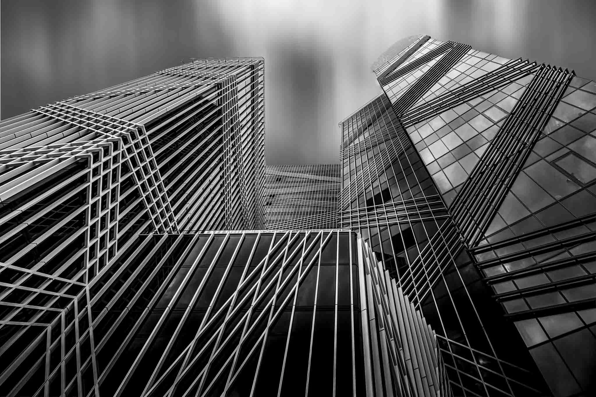 Black and white fine art architectural photograph looking upward between modern glass buildings, with strong geometric lines, converging perspectives, and motion-blurred clouds.