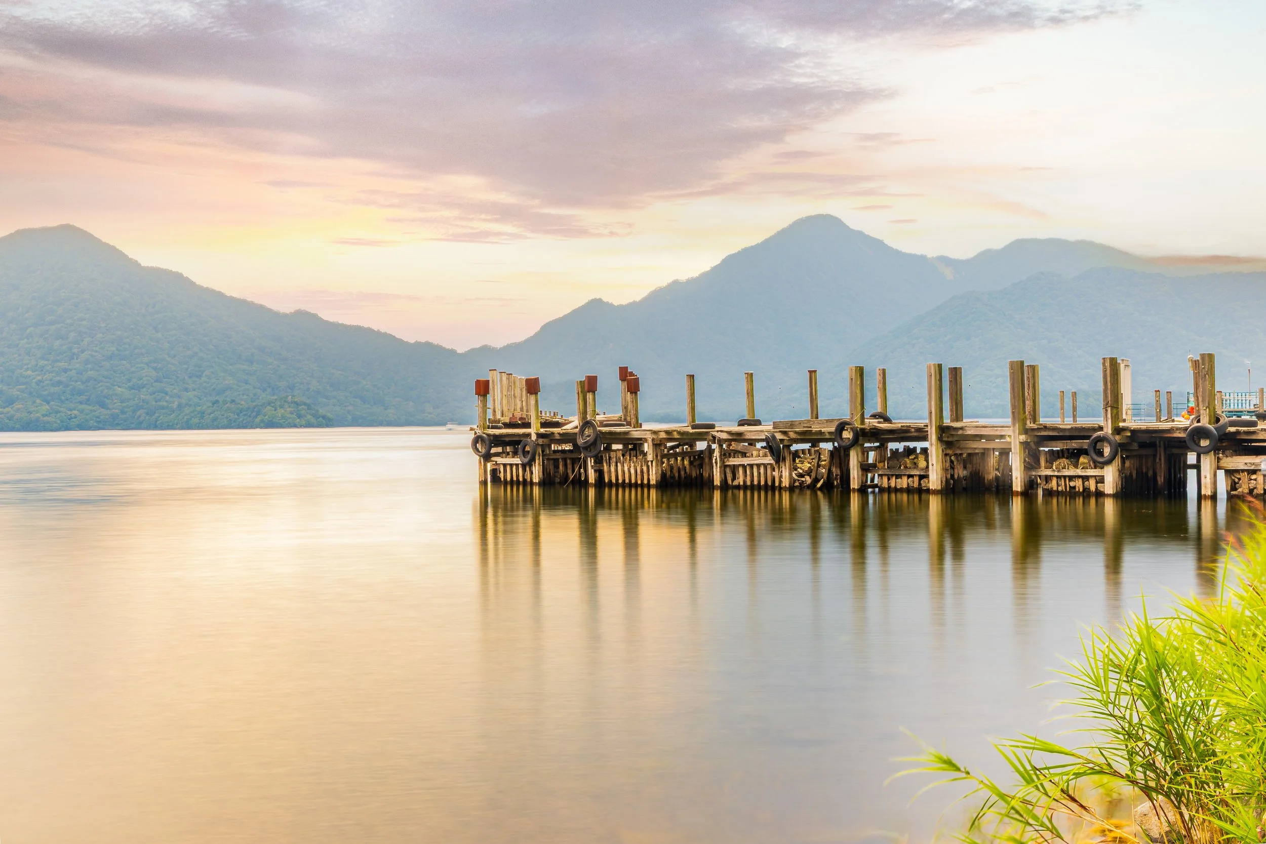 A wooden dock extending over a calm lake with mountains in the background during sunset.