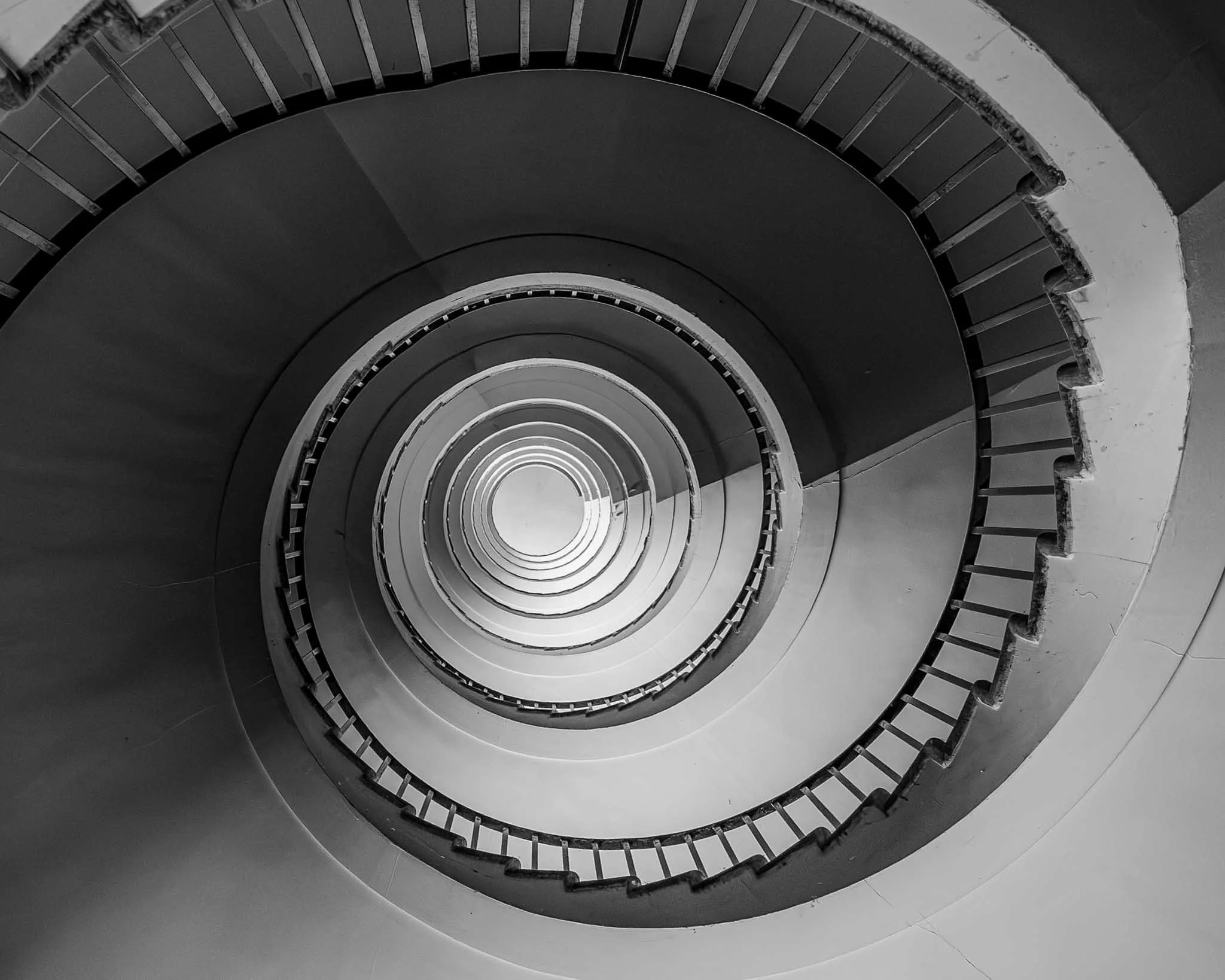 Black and white architectural photograph looking down a spiral staircase, with repeating curved lines leading toward a bright circular center.