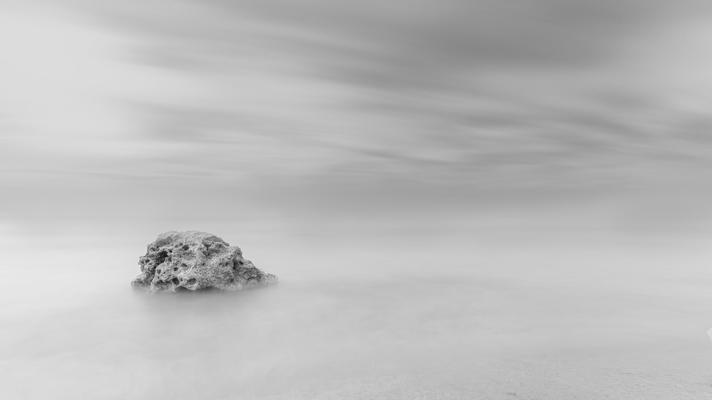 A black and white long exposure photograph of a sea with a single rock on the shore, smooth water, and blurred clouds in the sky.