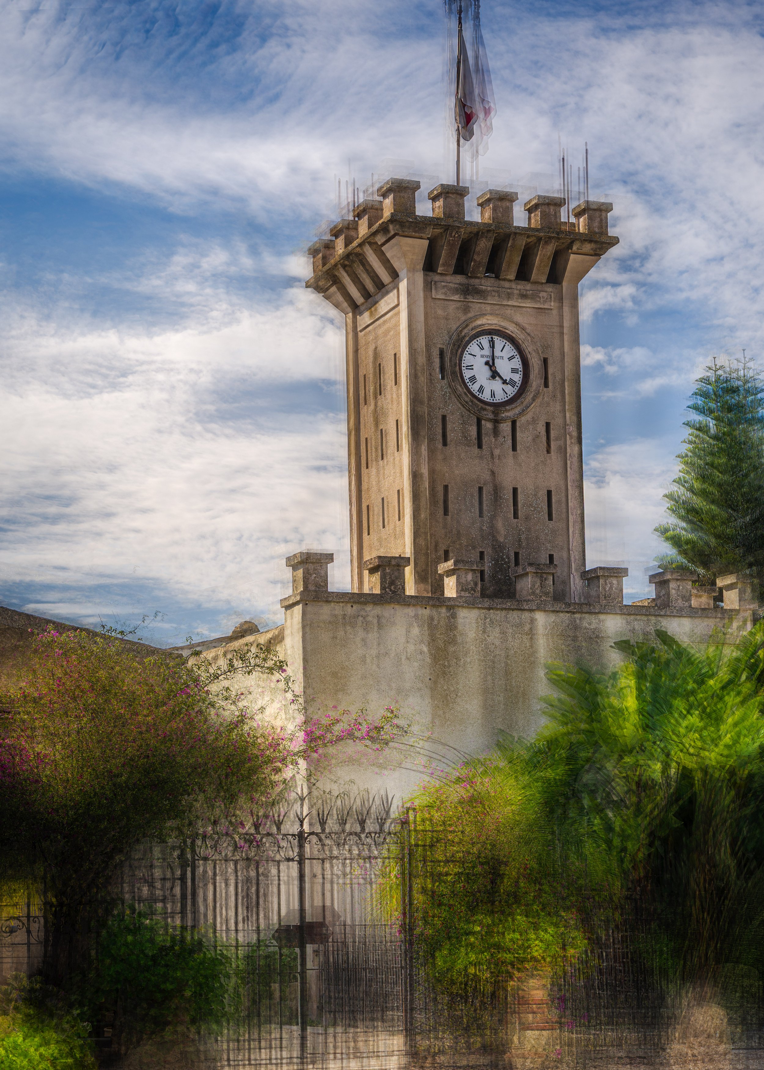 A tall stone clock tower with a clock face showing 2:35, topped with a flagpole, surrounded by greenery and a black wrought iron fence.