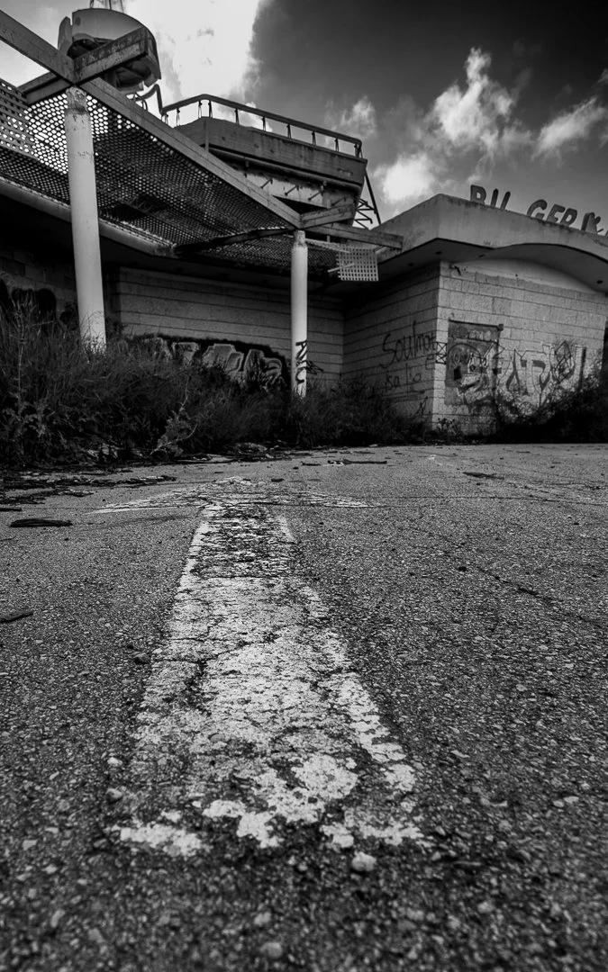 Black and white photo of an abandoned, cracked parking lot with overgrown weeds and graffiti on the building wall. A dilapidated structure is visible in the background against a cloudy sky.
