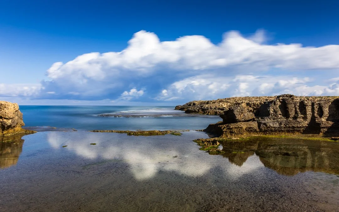 Coastal scene with rocks, calm water, and a blue sky with white clouds.