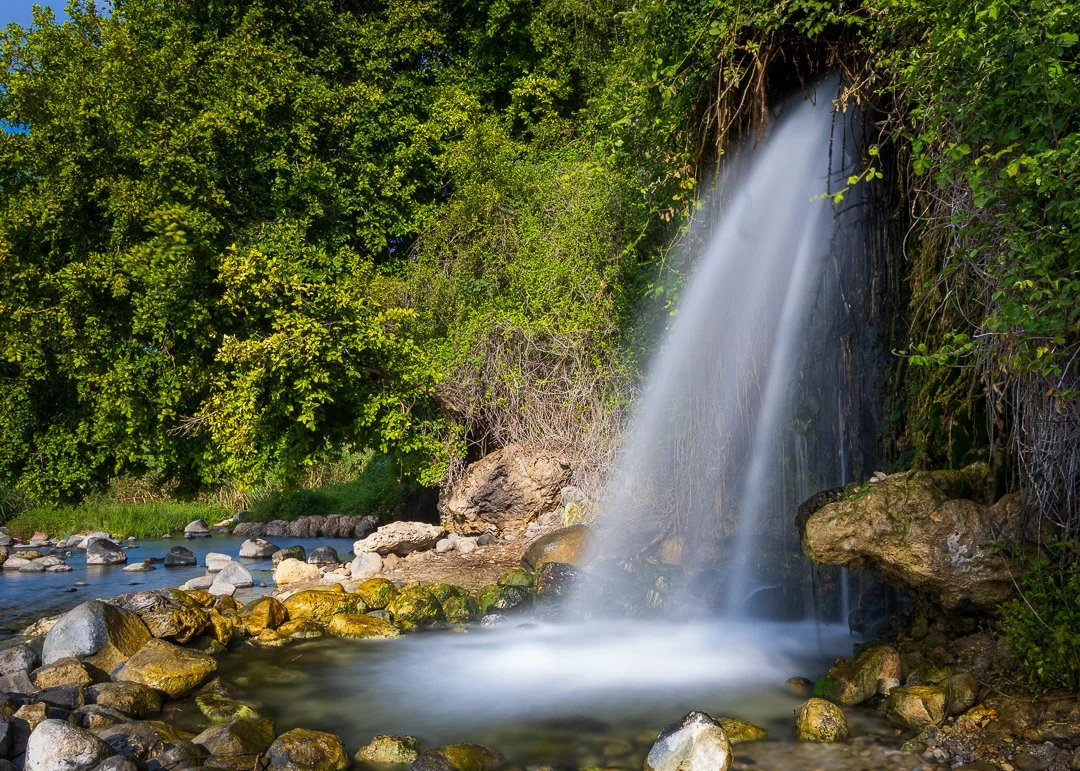 A waterfall flowing over rocks into a small pond, surrounded by lush green trees and foliage.