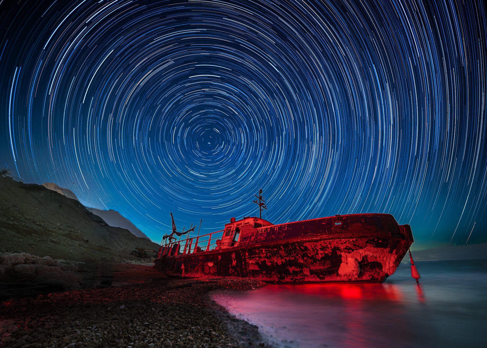 Star trail photograph over an abandoned red boat on rocky shore at night.