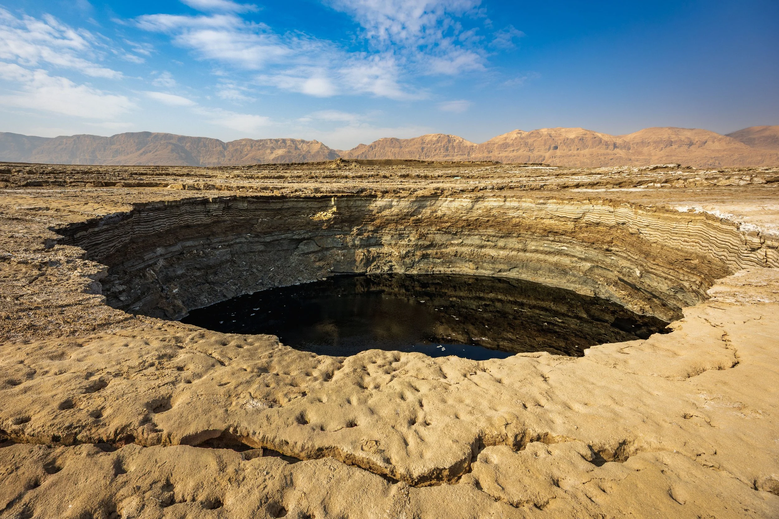 A large, deep sinkhole in a dry, desert landscape with layered rock formations surrounding it and mountains in the background under a partly cloudy sky.