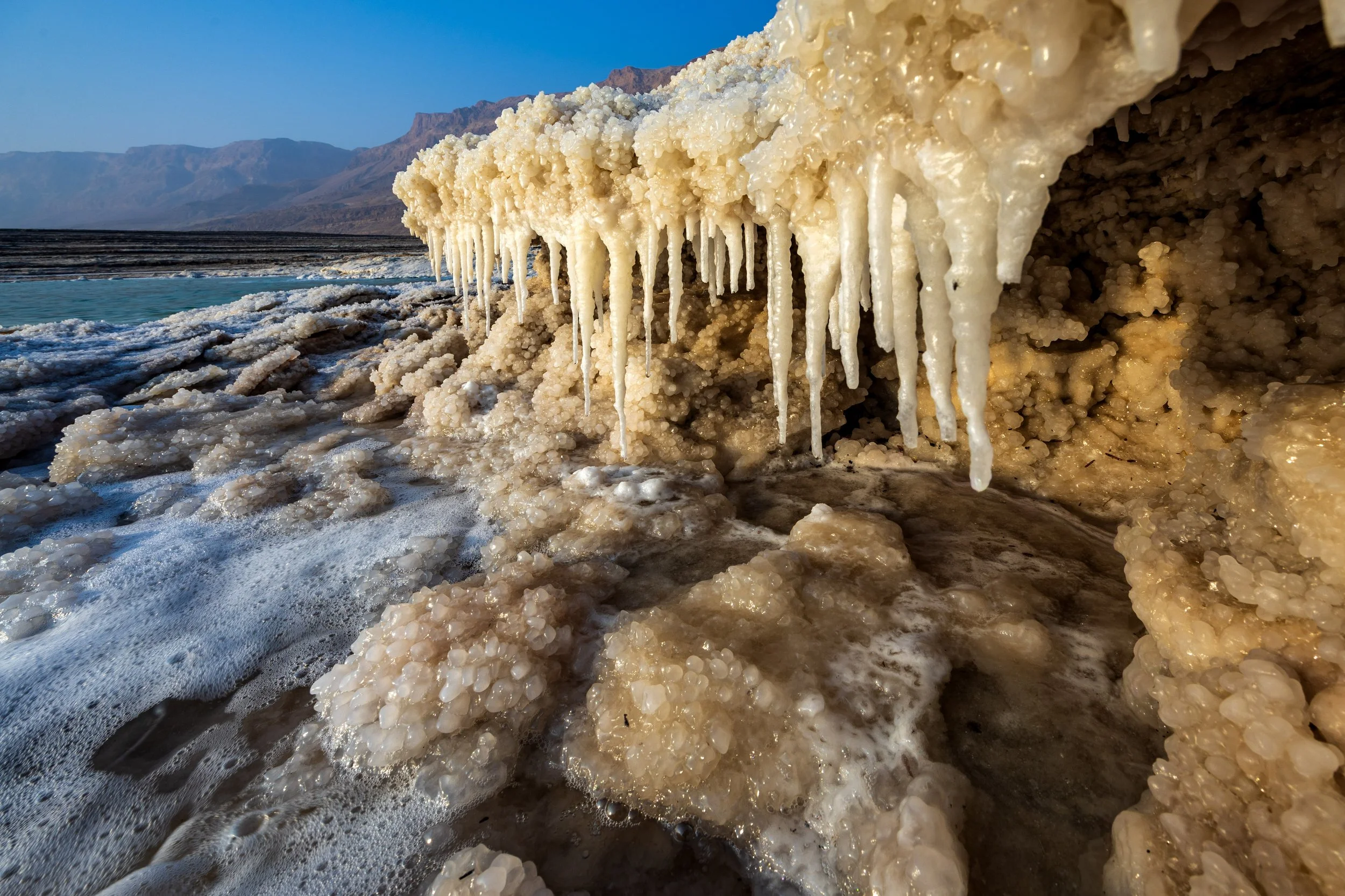 Salt formations with icicles along a shoreline with mountains in the background.