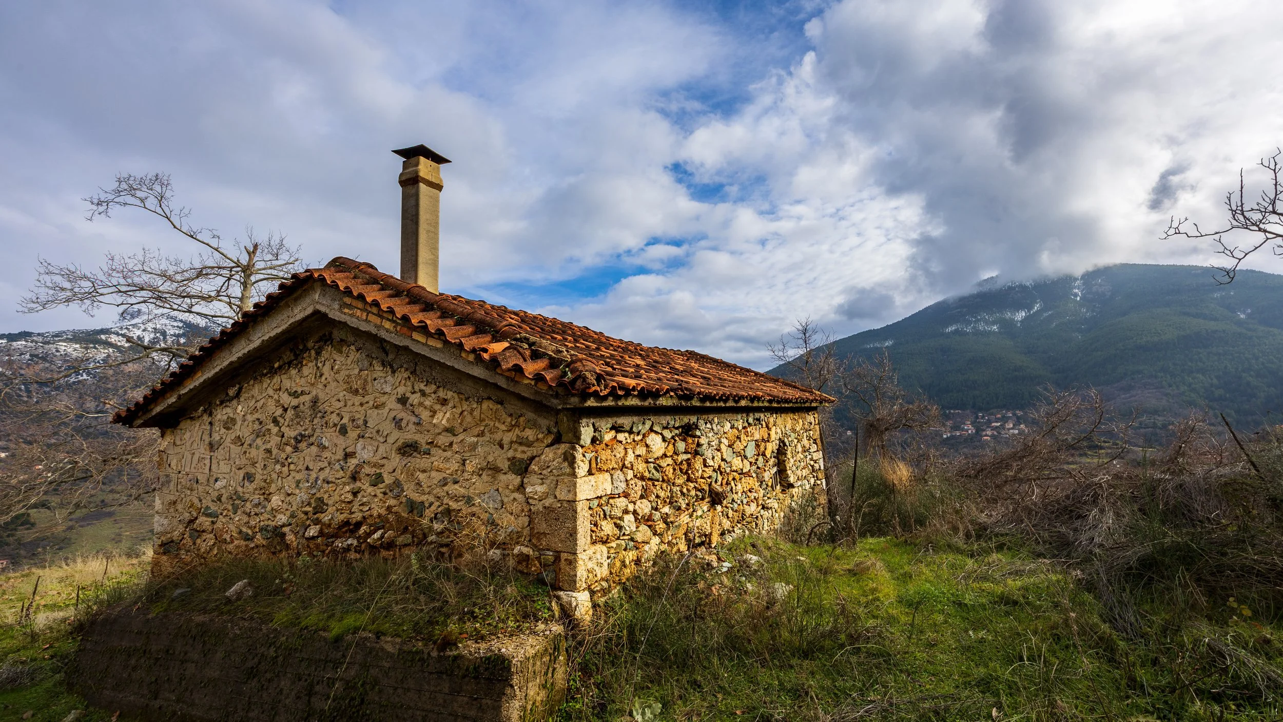 An old stone house with a red tiled roof and chimney in a rural landscape with mountains and trees, photograph by erez nudmanov