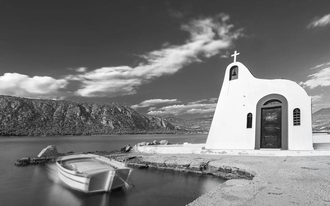 A small church with a cross on top, near the water, with a boat floating in the foreground and mountains in the background, in black and white.