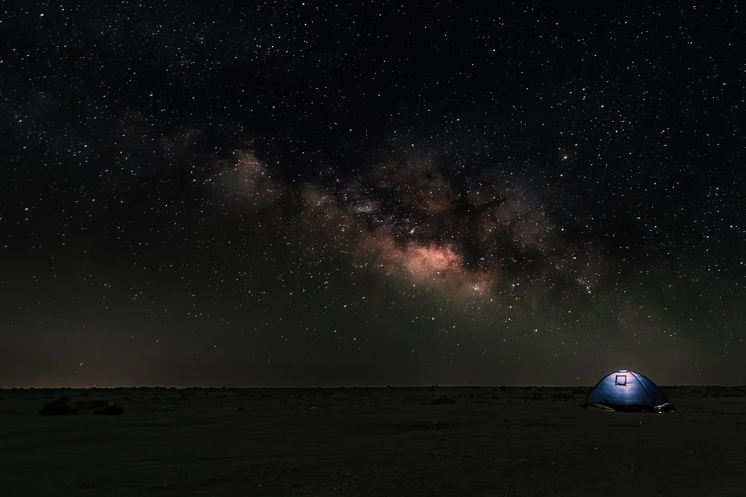 A night sky filled with stars and the Milky Way galaxy, with a small illuminated tent on the ground in the foreground.