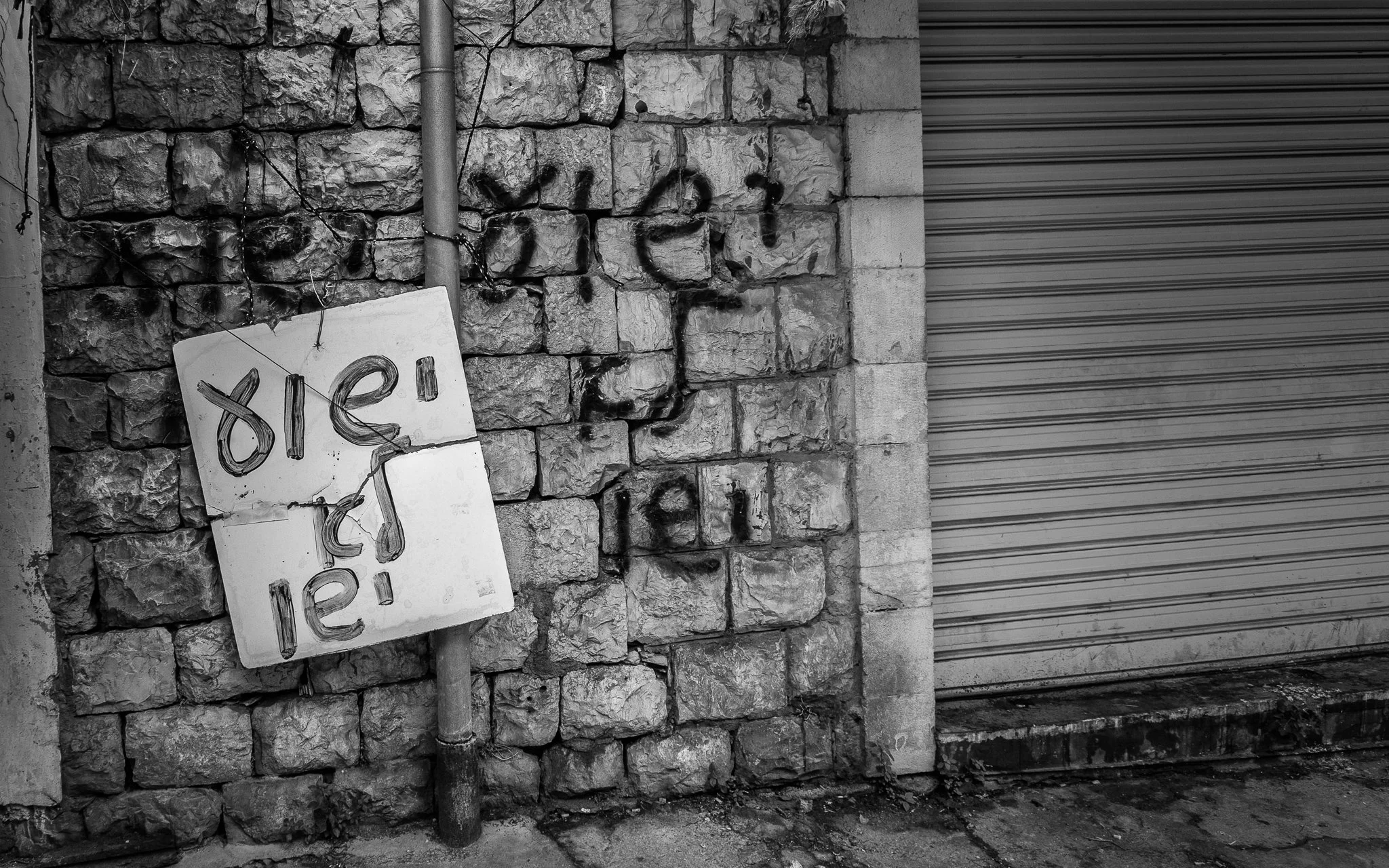 A black and white photo of a stone wall with graffiti, a metal pipe, and a sign that reads 'bief!' hanging from the pipe. A closed metal roll-up door is on the right side.