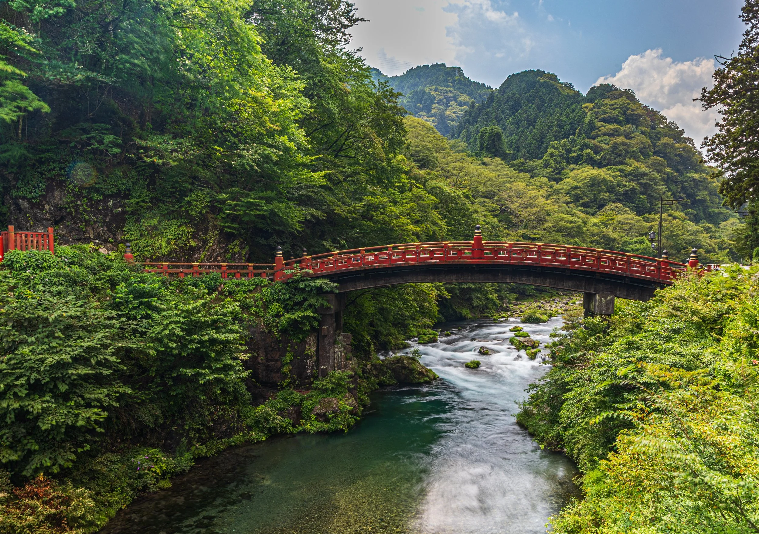 A red bridge spans a river surrounded by lush green trees and hills in a scenic landscape.