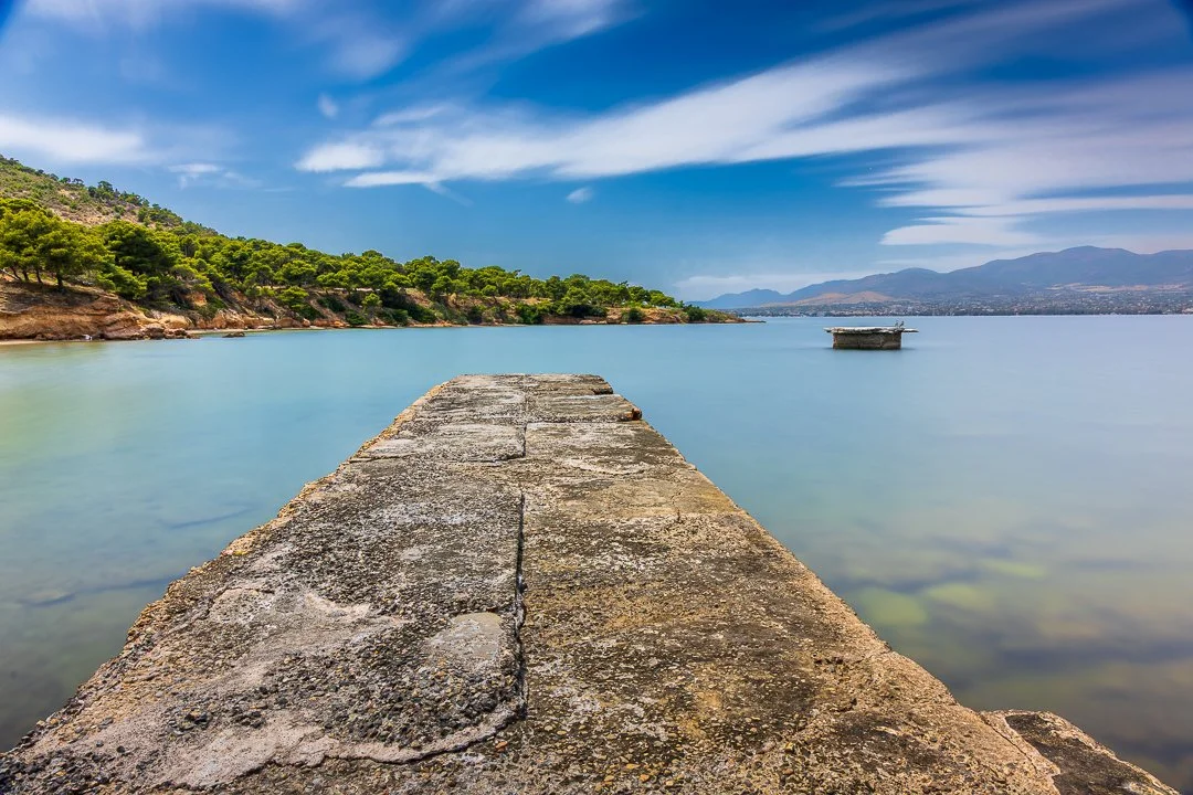 A stone pier extends into a calm lake, with green trees on the left, distant mountains on the right, and a partly cloudy blue sky overhead.