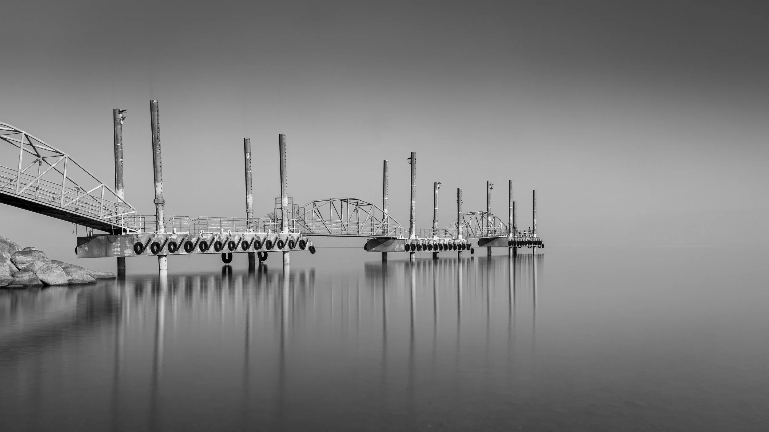 A black and white fine art photo of an empty pier extending into calm water, with rocks on the left side and a clear sky photography by erez nudmanov