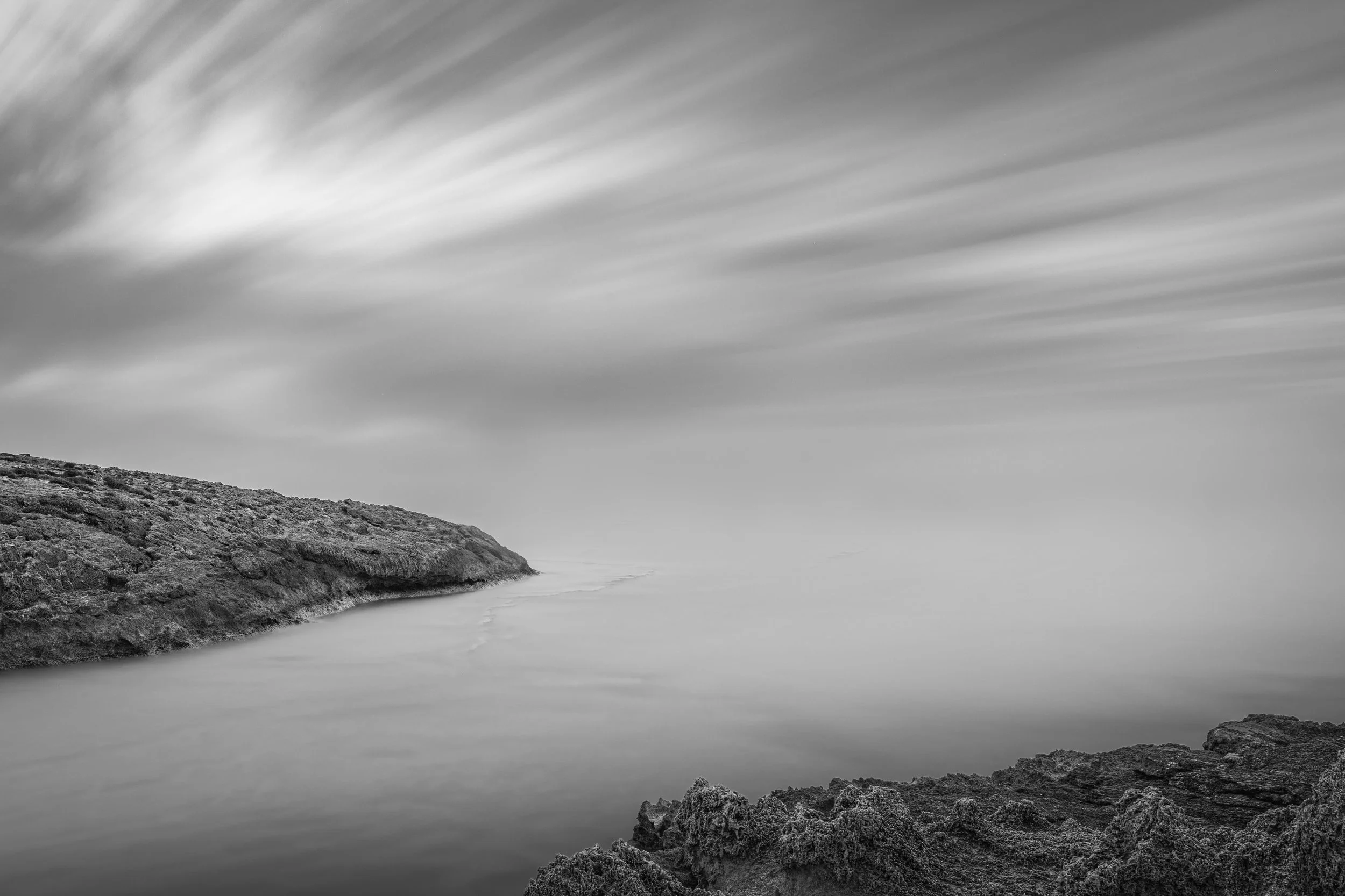 Black and white long exposure photograph of a rocky coastal landscape with water and sky in the background.