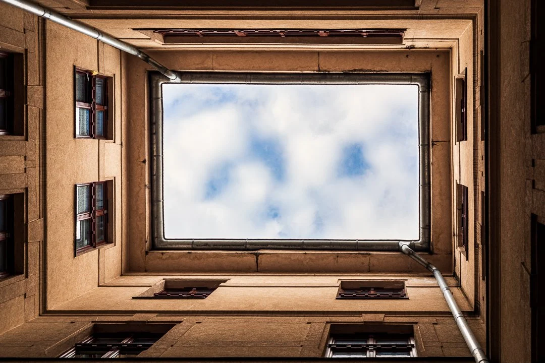 Looking up from the courtyard of a building, showing the sky through a rectangular opening at the top, surrounded by beige walls and windows with brown frames.