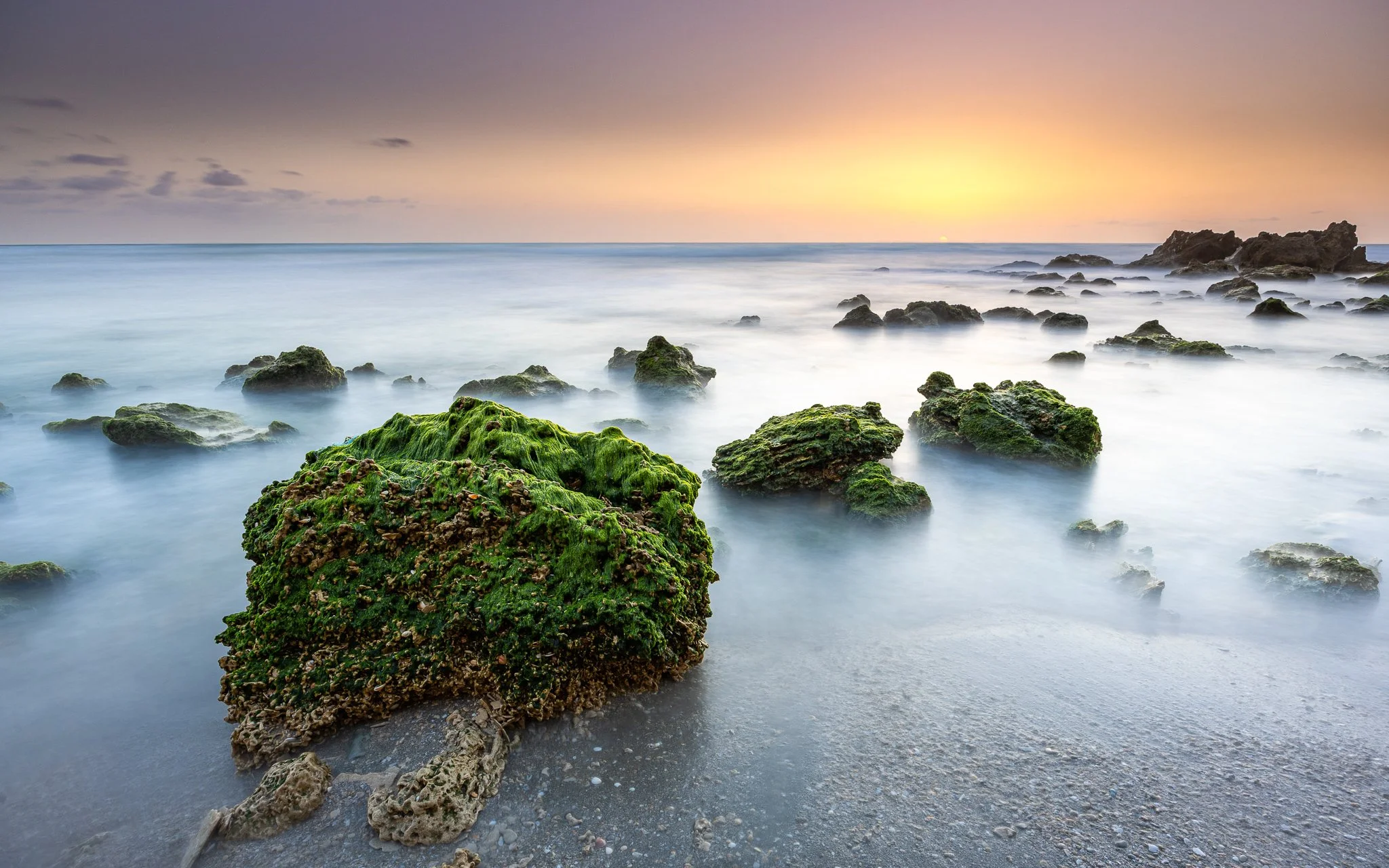 Sunset over a beach with moss-covered rocks in the tide, creating a serene seascape.