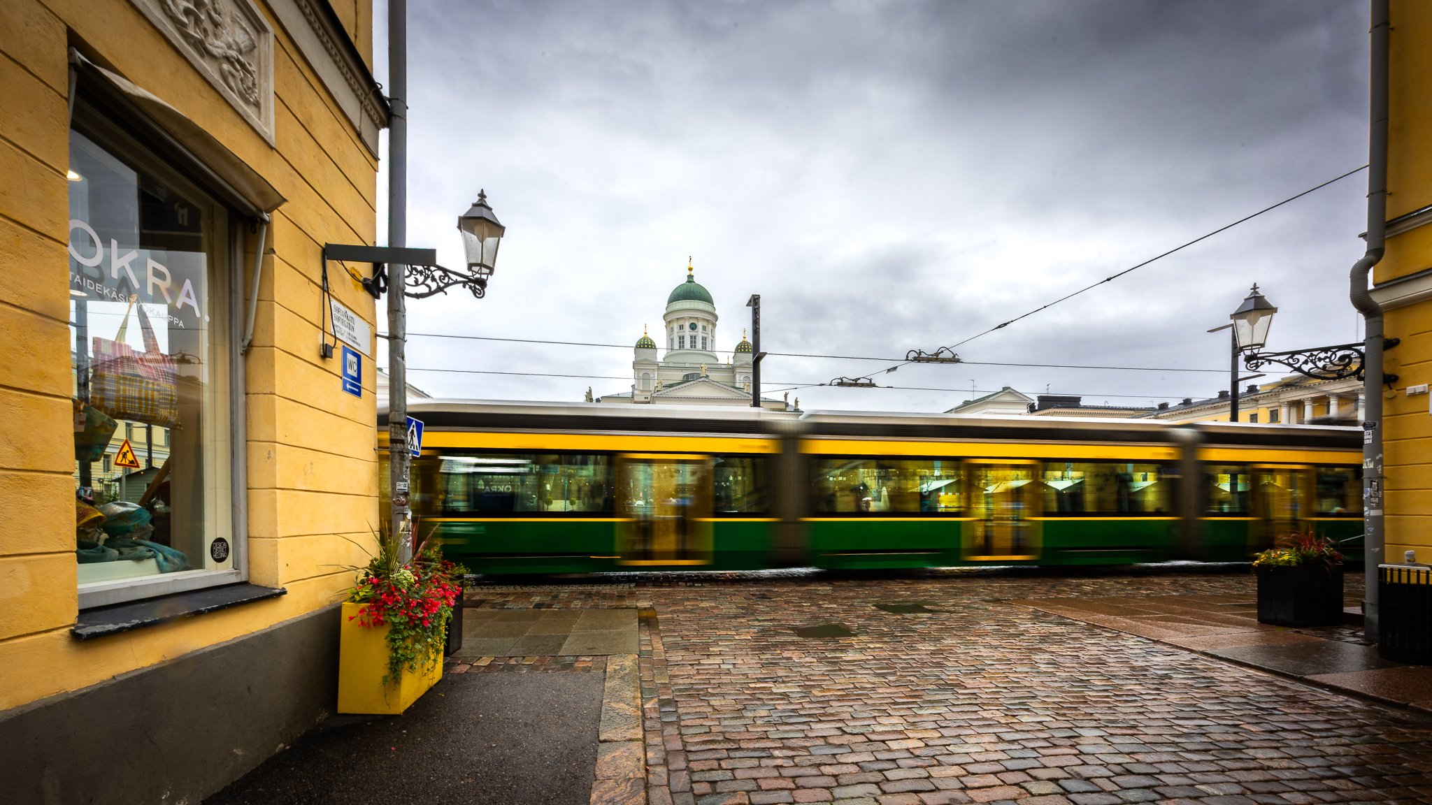 A city street with a yellow building on the left, a green and yellow tram passing by, and a domed church with green roofs and golden accents in the background. The sky is cloudy.