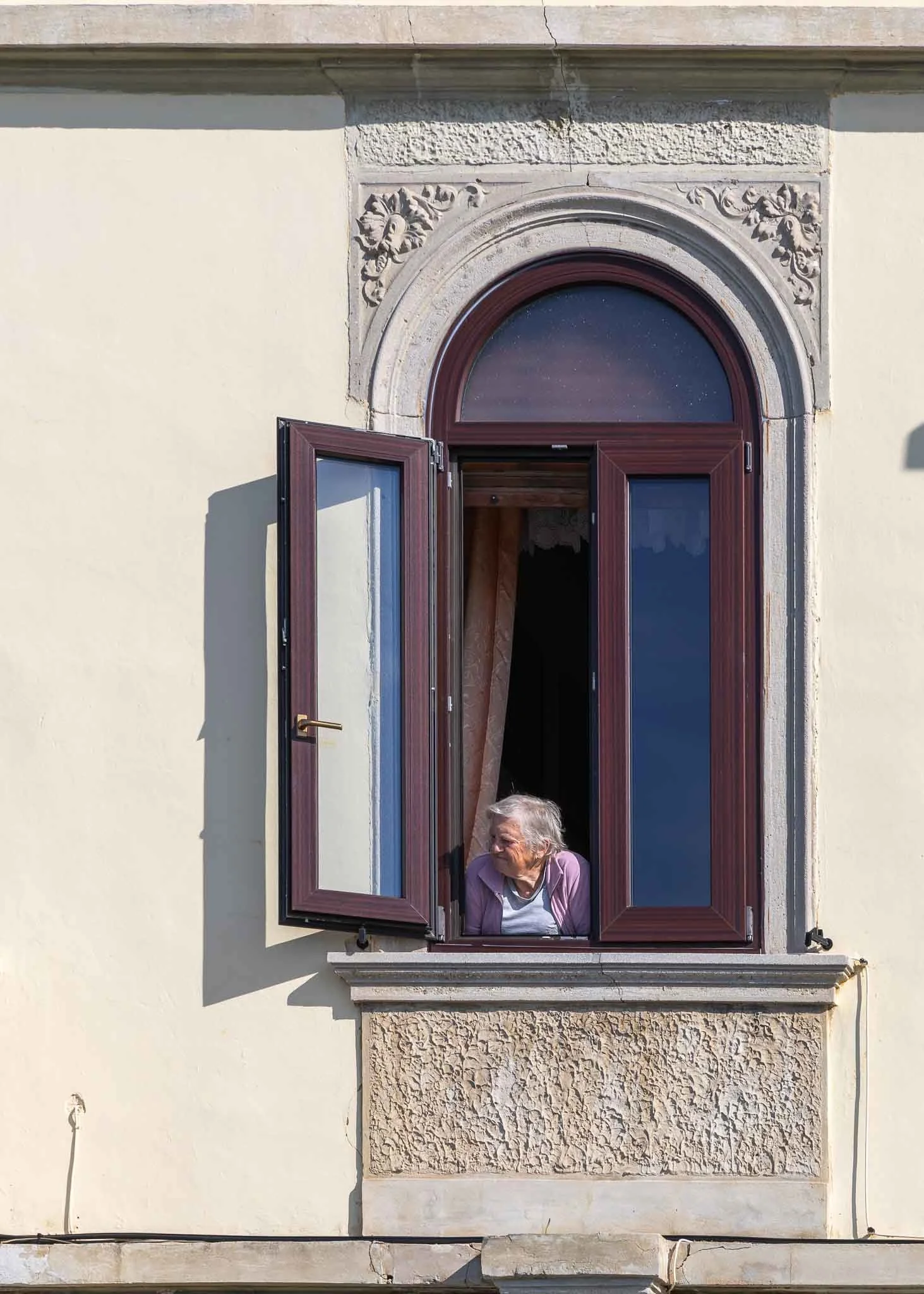 An elderly woman leaning out of an ornate arched window with wooden shutters, looking to the left in warm sunlight against a cream-colored historic building facade in Slovenia.