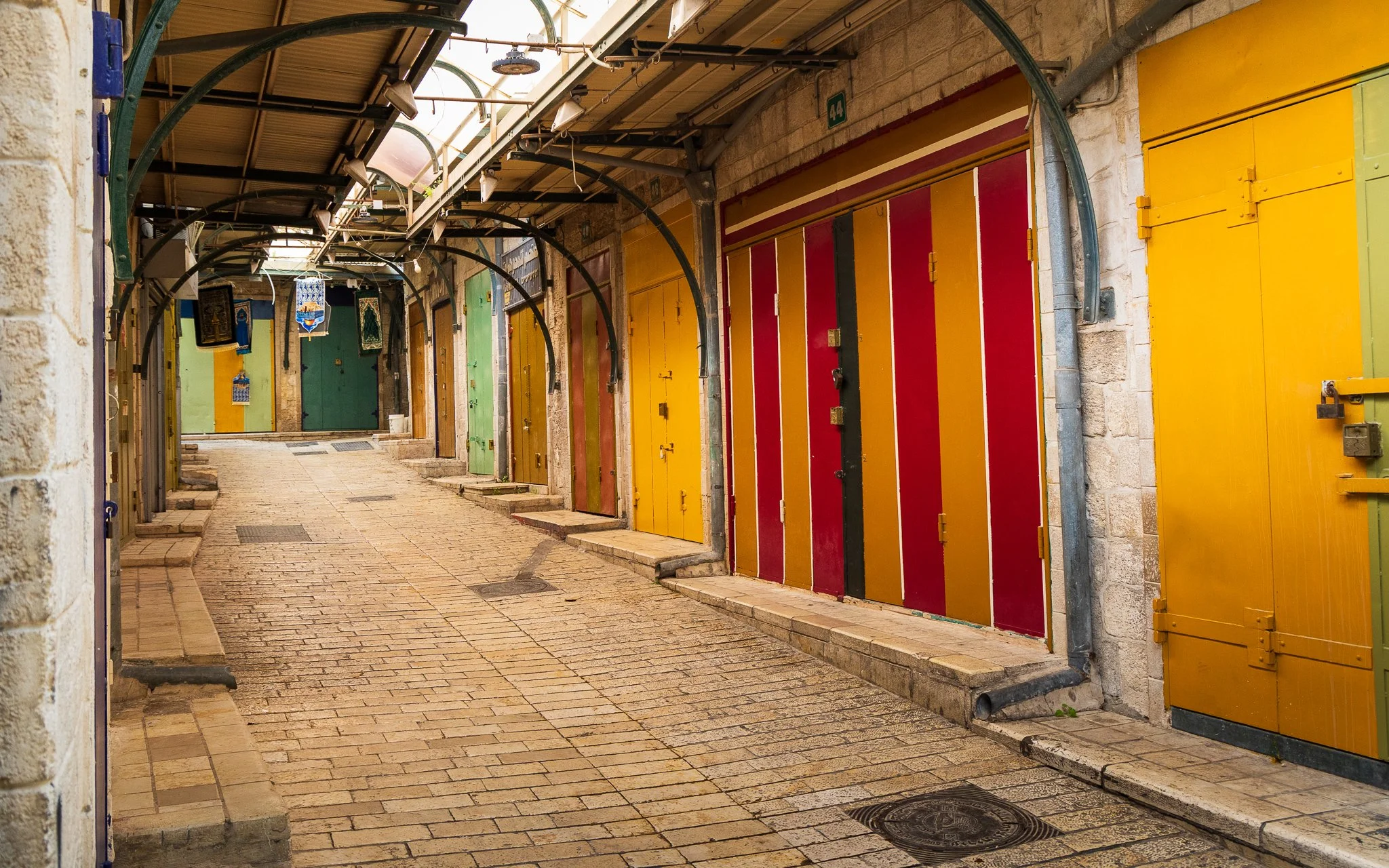 A cobblestone alleyway with colorful closed doors painted in red, yellow, green, and black, and covered walkways with metal arches overhead.