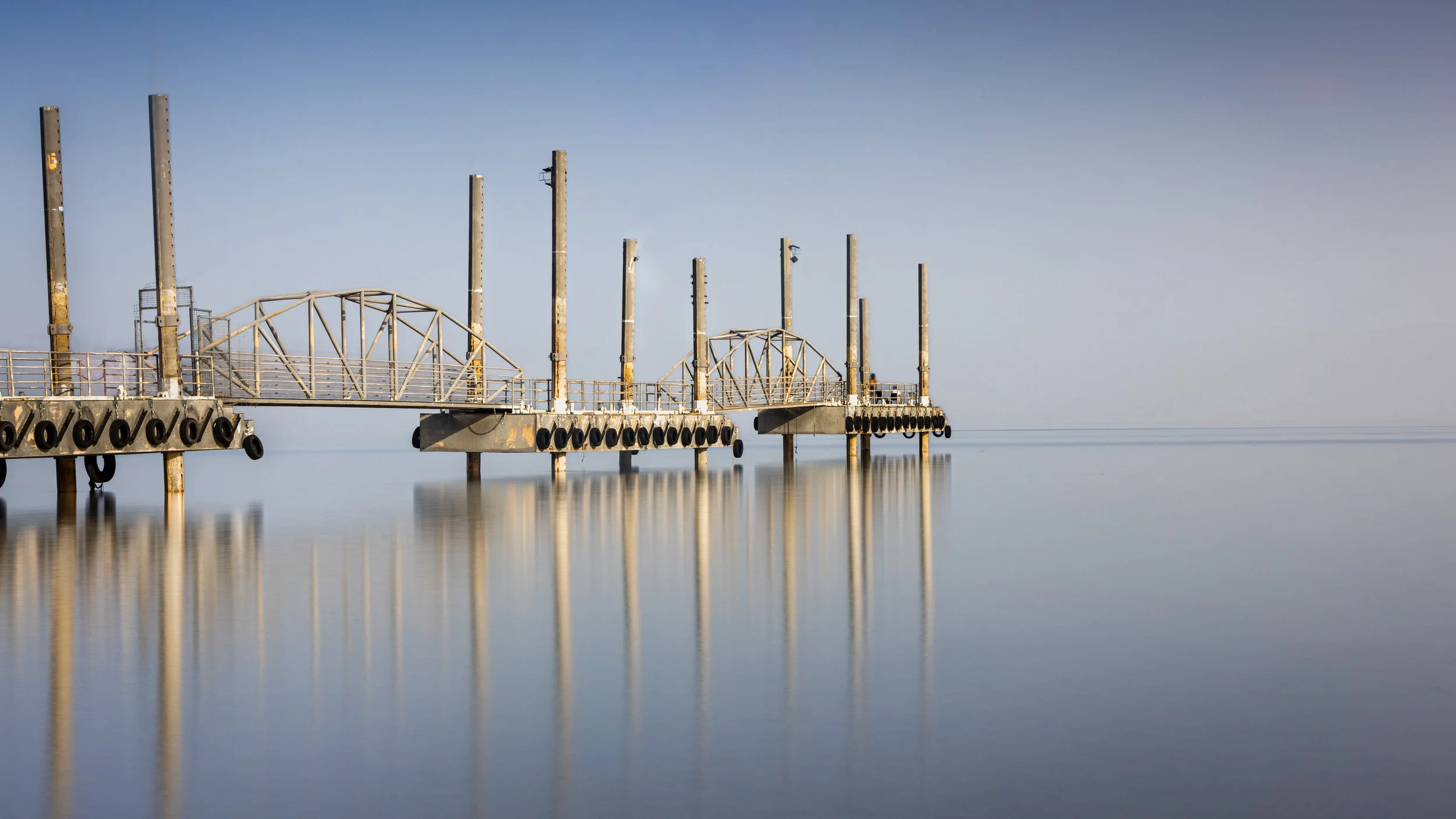 fine art pier extending into calm water with reflections, under a clear blue sky photography by erez nudmanov