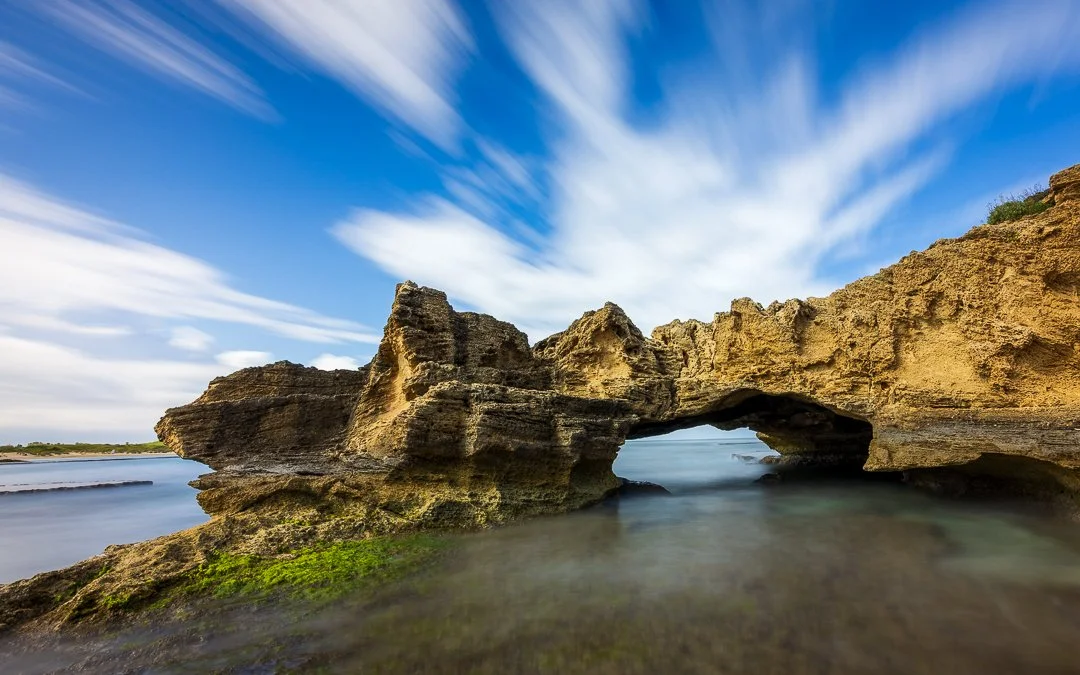 A natural rock formation with a large archway in the middle, surrounded by water and a partly cloudy sky.