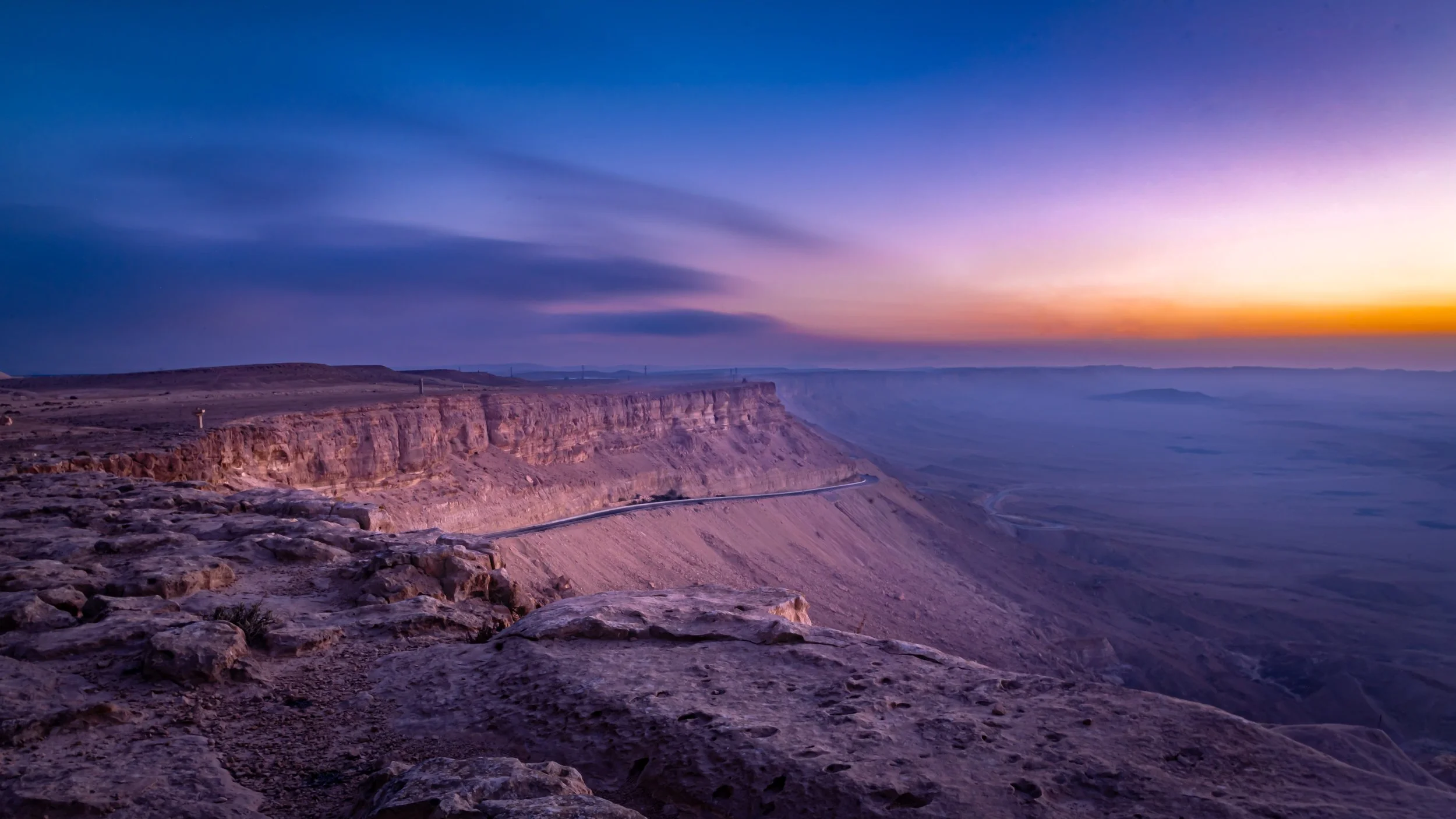 A desert canyon landscape at sunset with purple and orange sky, rocky terrain in the foreground, and a winding road along the canyon edge.