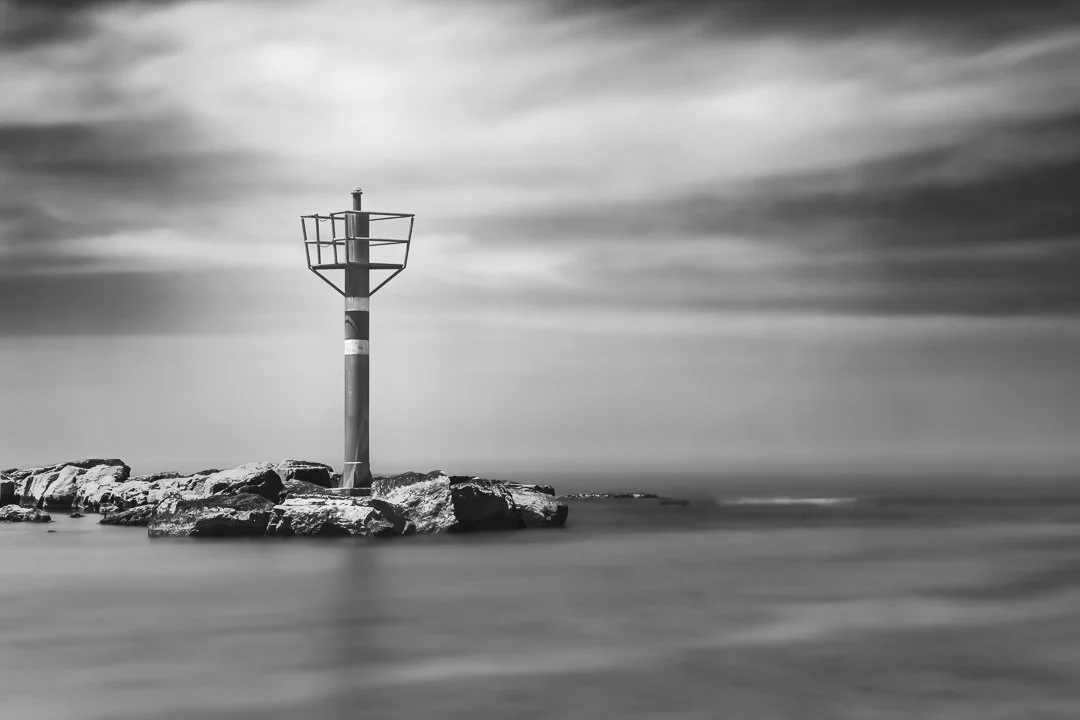 A black and white photo of a small lighthouse or navigation tower on rocks in the water with a cloudy sky in the background.