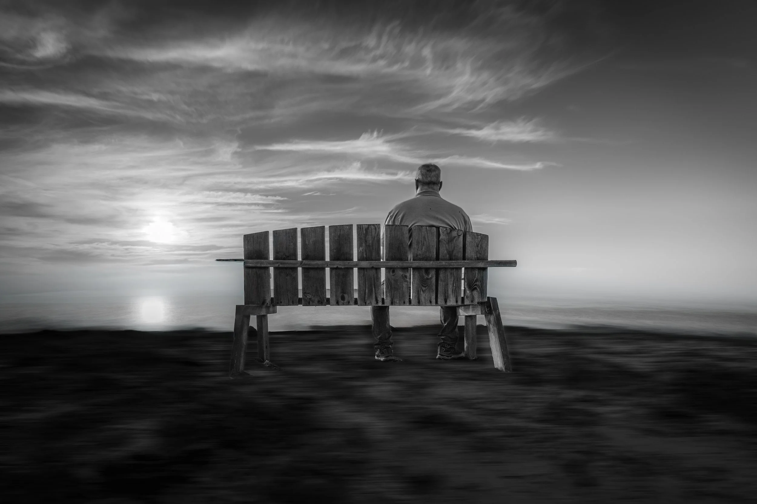 A solitary man sitting on a wooden bench facing the ocean during sunset or sunrise, with a cloudy sky above, in black and white.