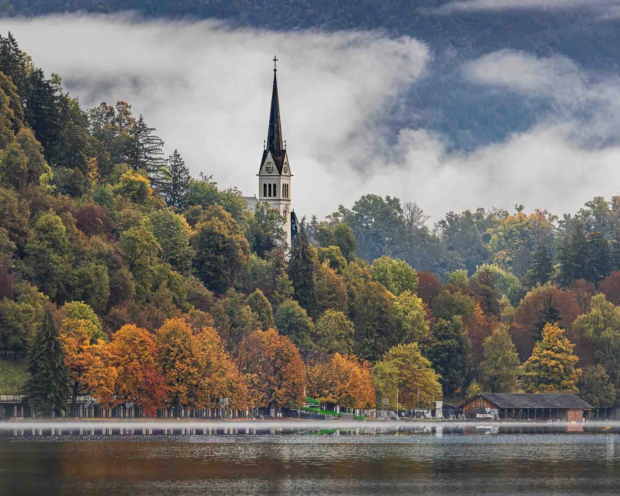 Bell tower of the Church of the Assumption on Lake Bled island, surrounded by autumn trees, mist and dramatic clouds in Slovenia.