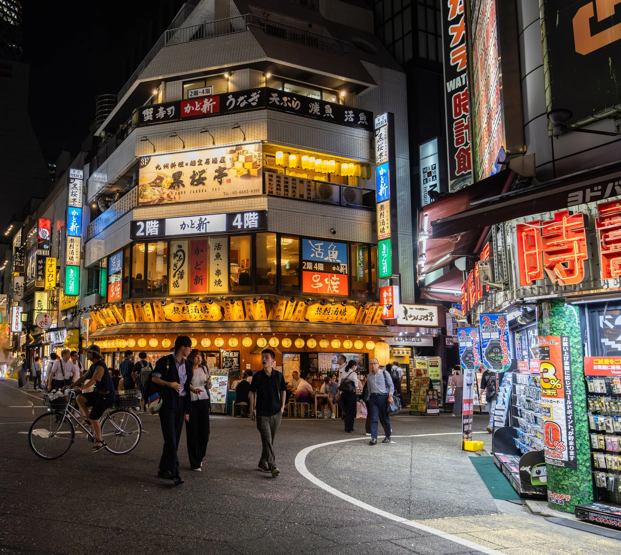Night scene of a busy city street in Japan with colorful neon signs and people walking, some with bicycles, and a restaurant with illuminated lanterns.