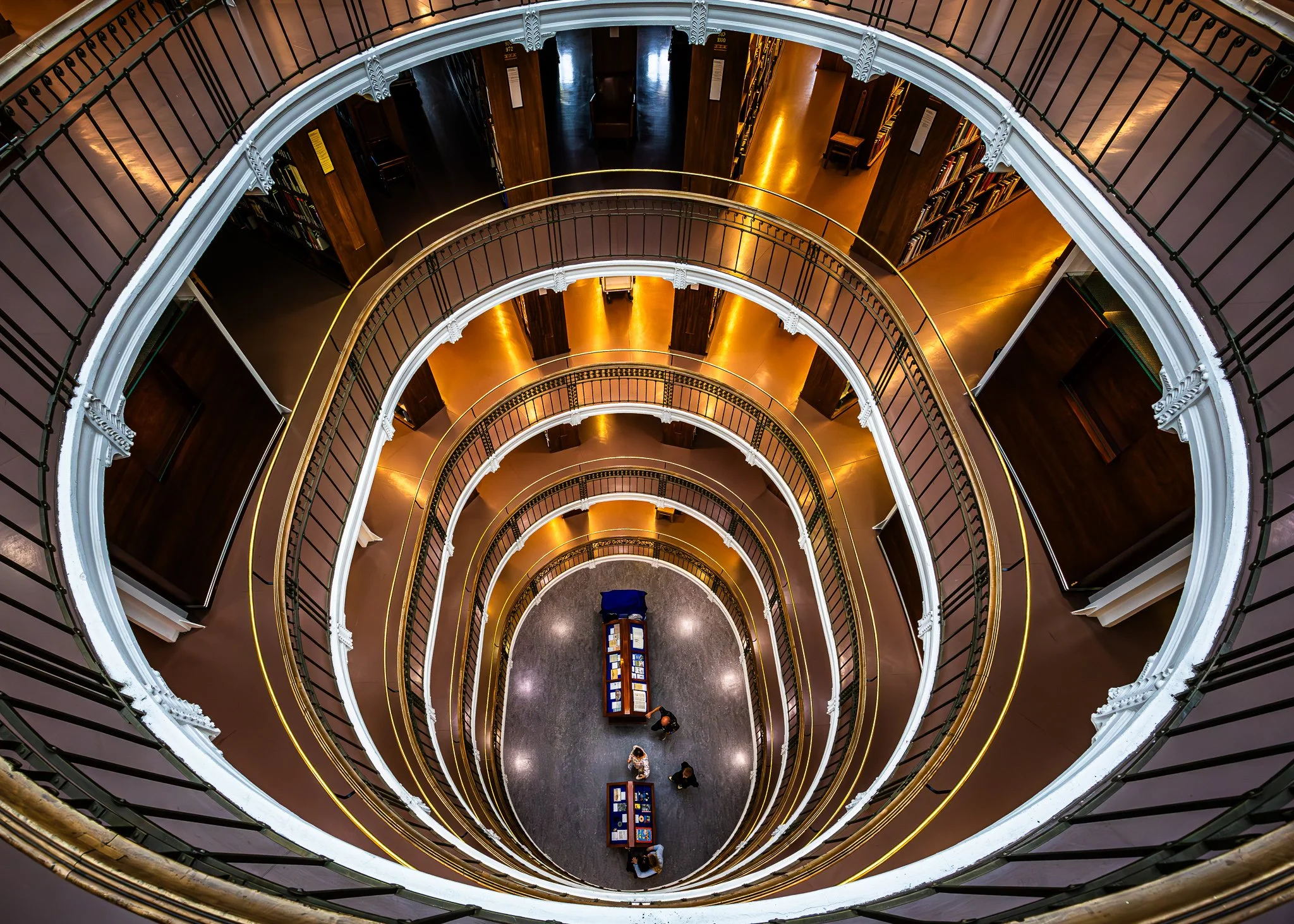 View from the top of a spiral staircase in a library, showing multiple levels with bookshelves and a few people on the bottom floor.