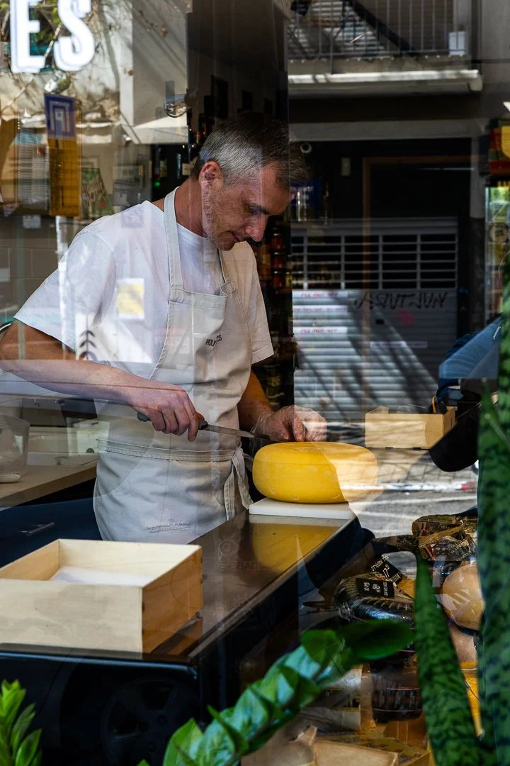 A man in a white apron slicing cheese behind a glass window in a shop.