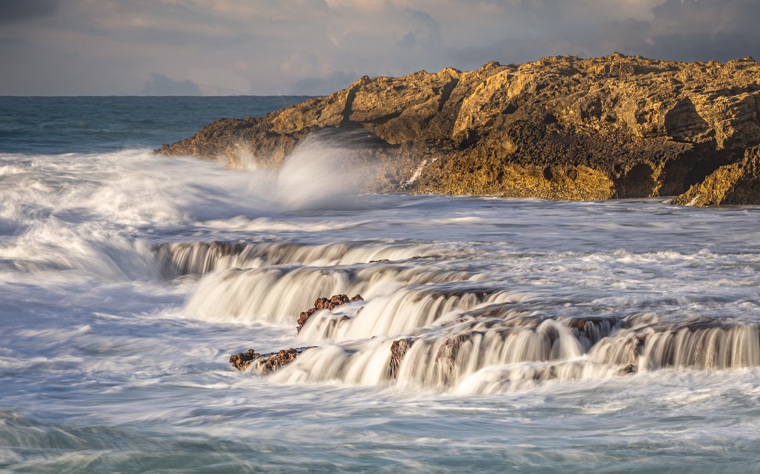 Waves crashing against a rocky shoreline under a cloudy sky at sunset.