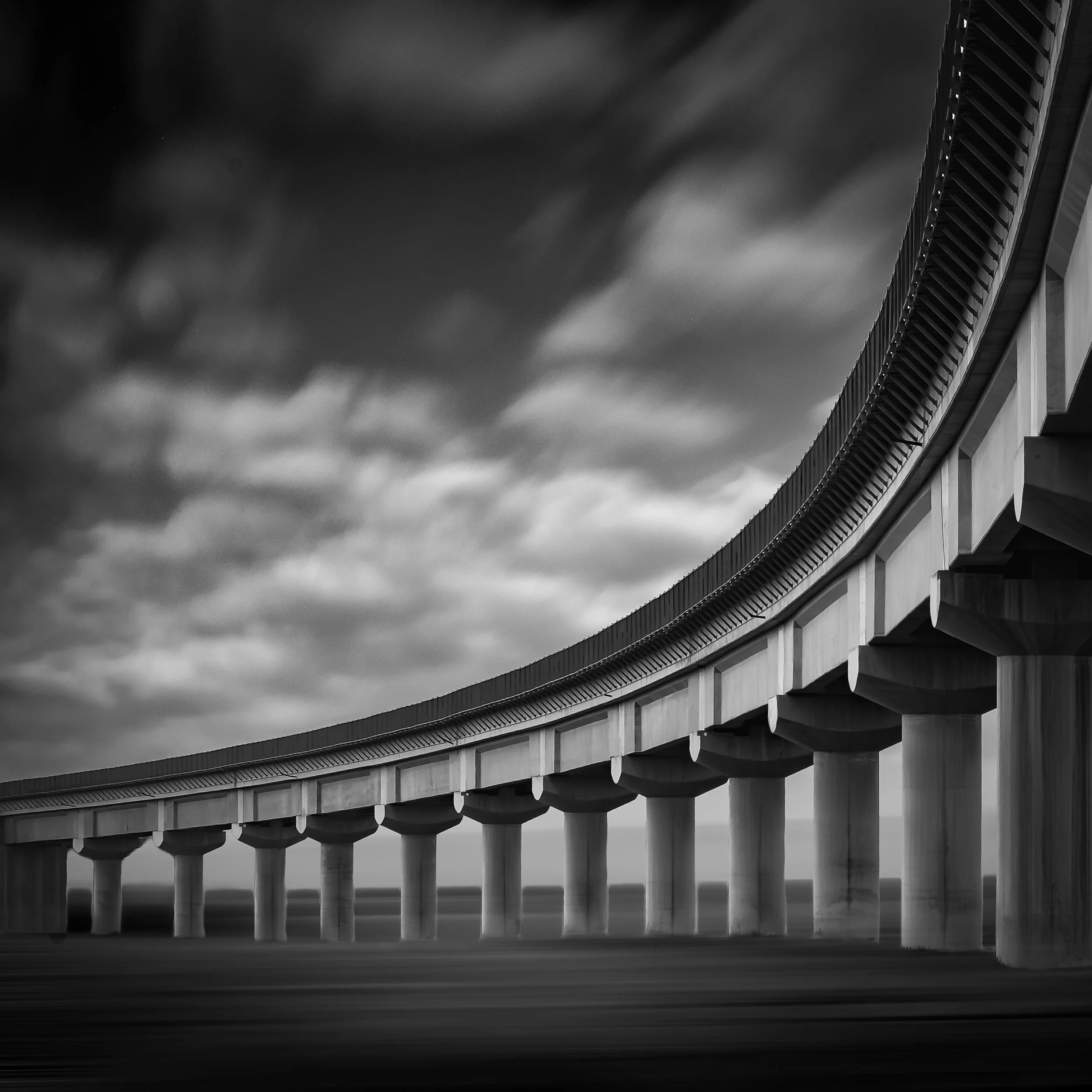 A black and white photograph of a long bridge with multiple supporting pillars, arching over open space with a cloudy sky above.
