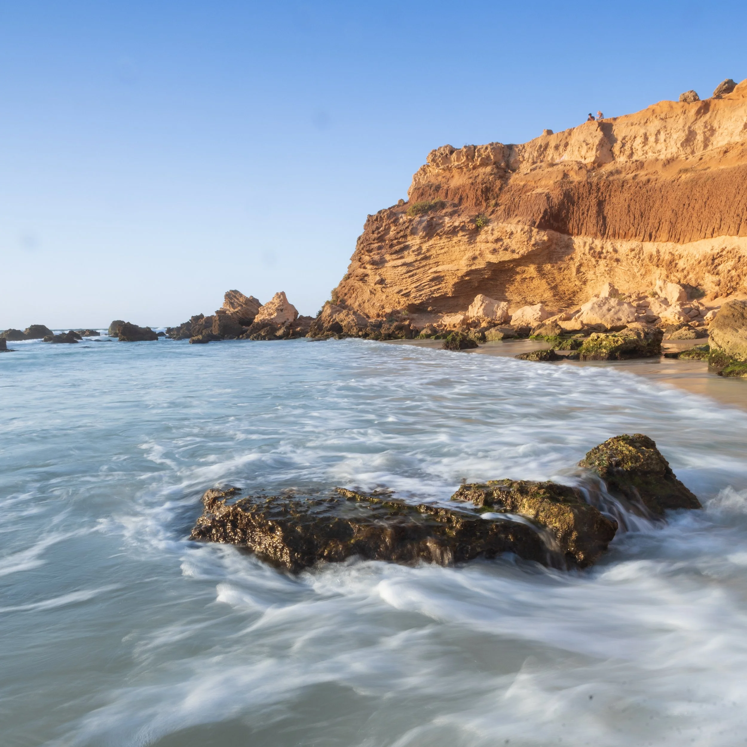 Sunlit rocky cliff overlooking the ocean with waves crashing against the rocks and sandy beach below.