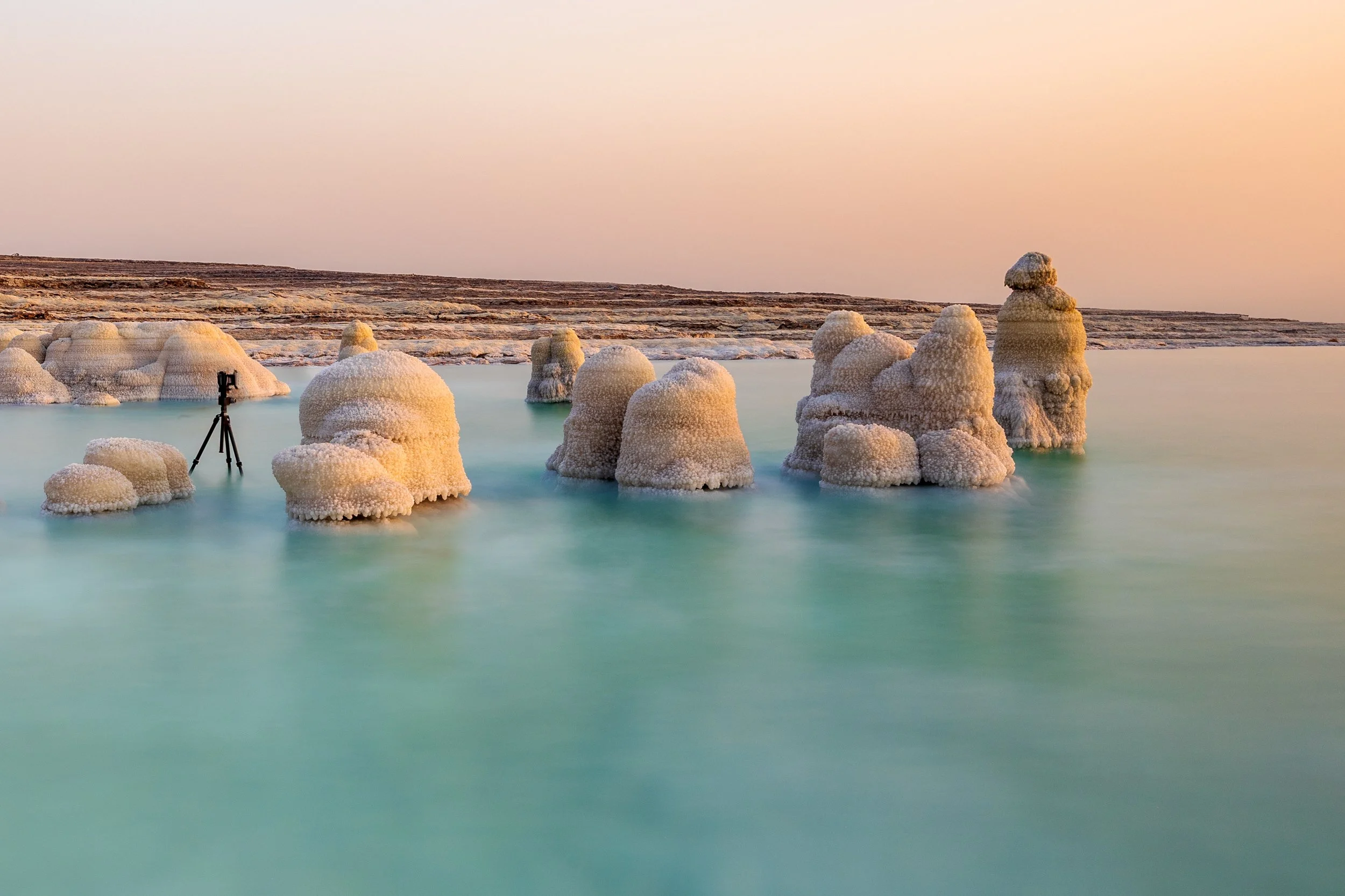 Salt formations emerging from a turquoise body of water, with a camera on a tripod nearby, under a pastel-colored sky.