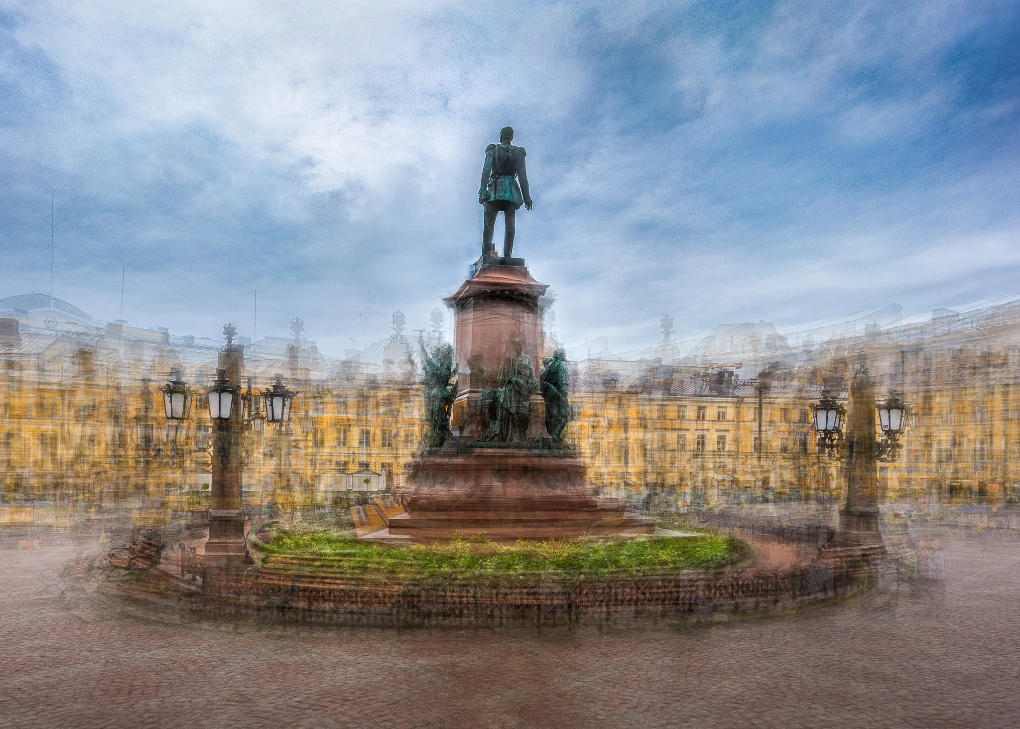 A statue of a man stands on a pedestal, surrounded by ornate lamp posts, with a historic cityscape and blue sky with clouds in the background.