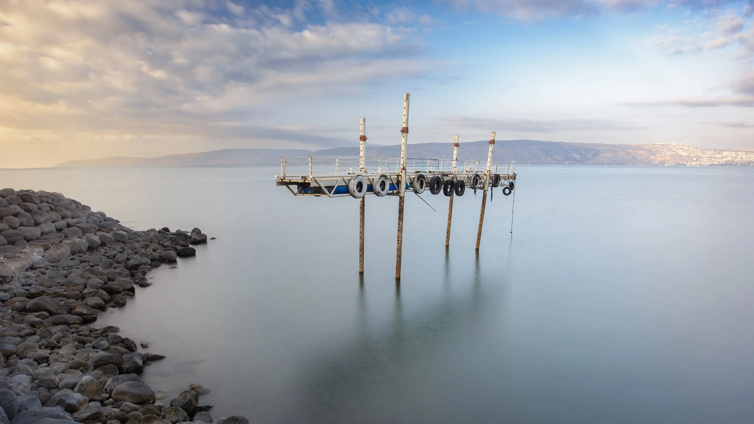 An old, unused floating dock or platform with tires hanging from its sides, partially submerged and tilted in the water near a rocky shoreline, with a hilly landscape and a cloudy sky in the background.