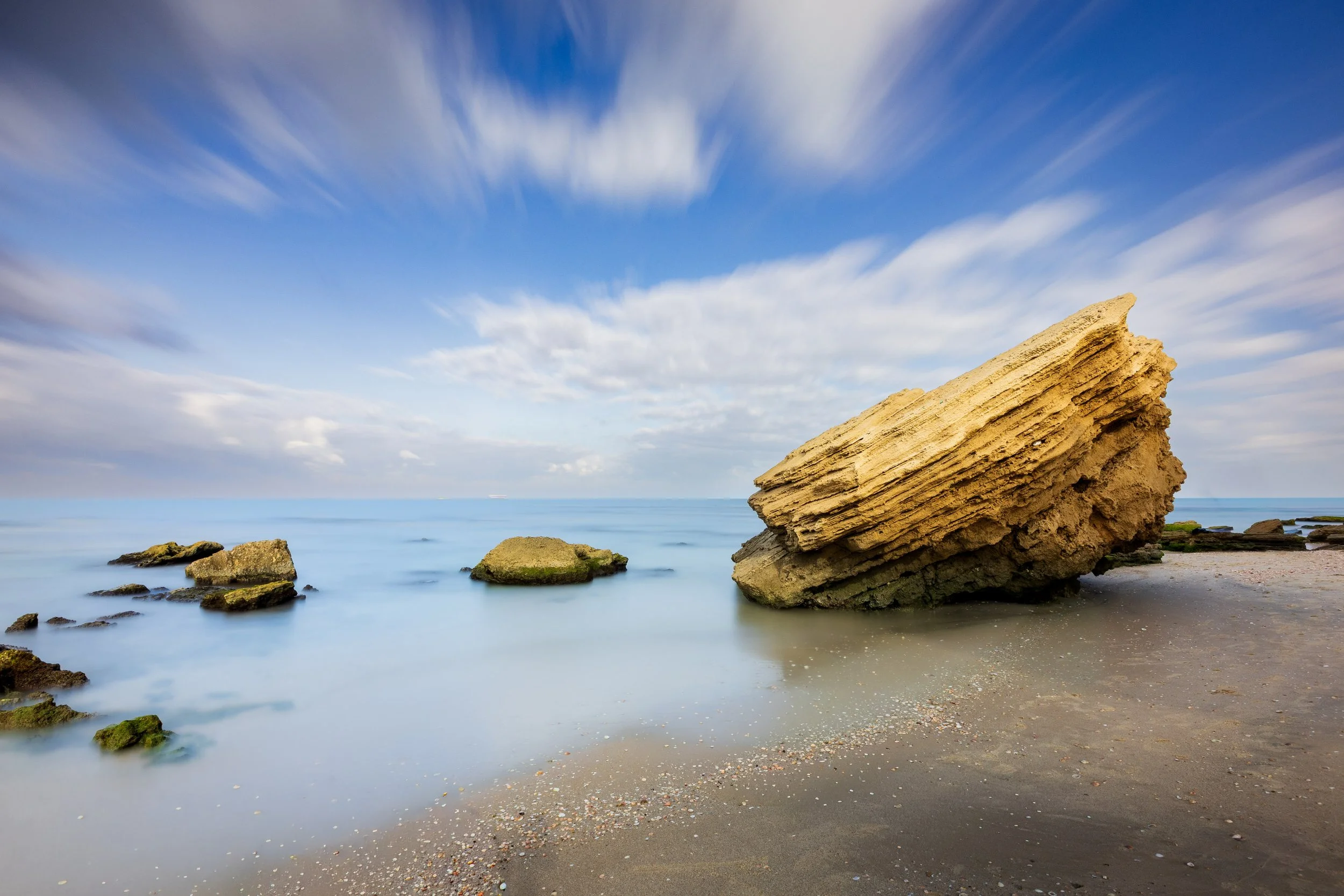 A large tilted rock on a sandy beach with smaller rocks nearby, calm ocean water, and a partly cloudy blue sky.