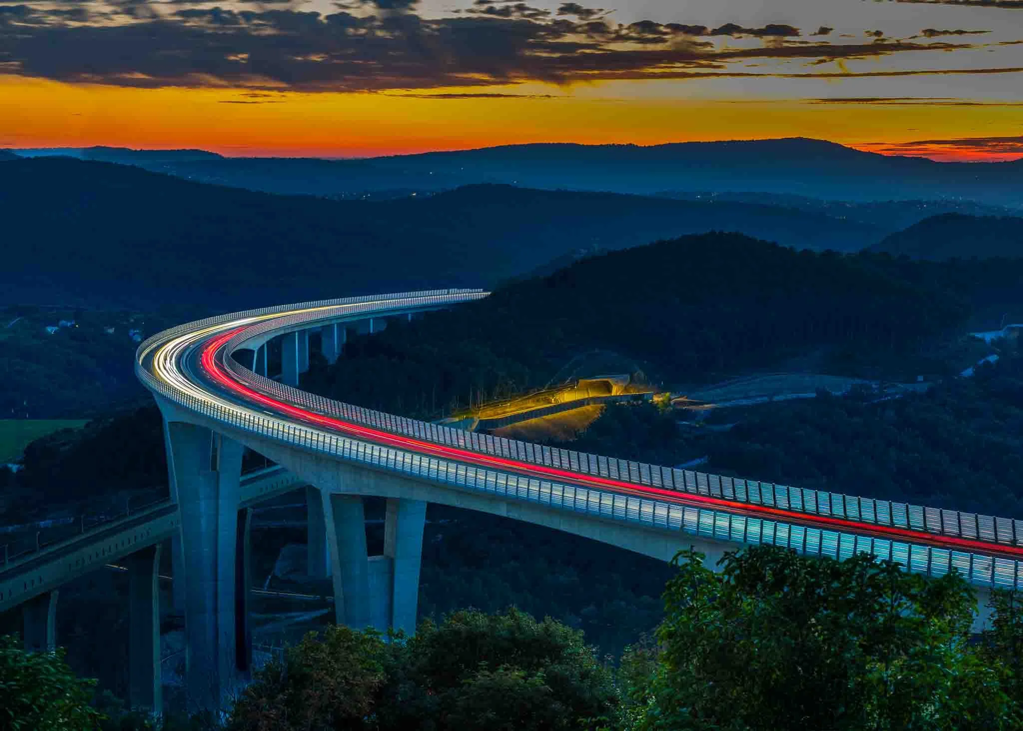 Long exposure light trails curving along a high concrete viaduct above forested hills beneath a vivid orange sunset sky in Slovenia.