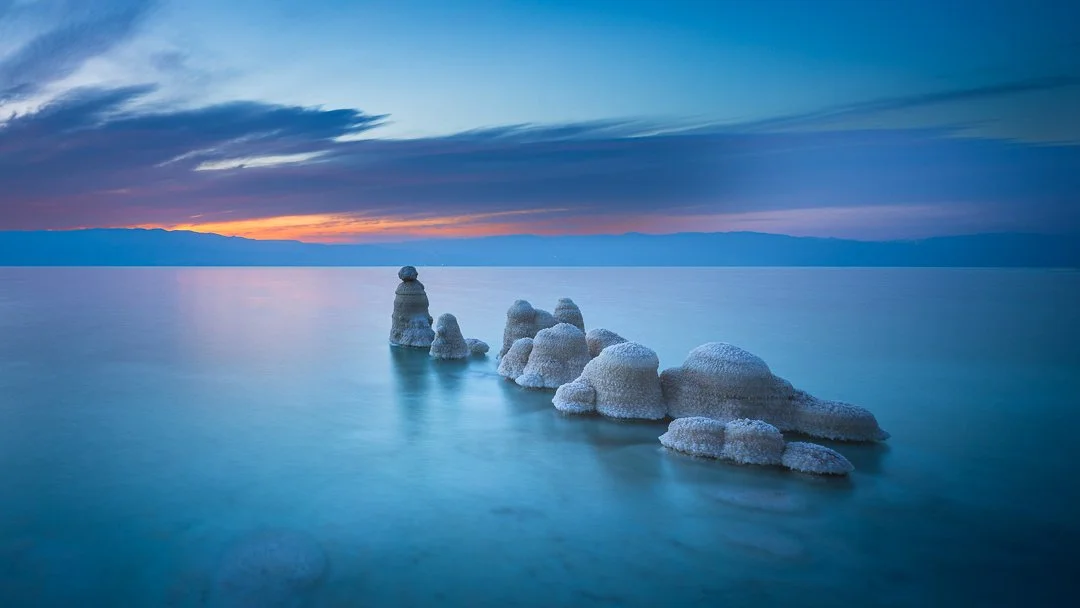 Salt formations emerging from the water at sunset with a colorful sky and mountains in the background.