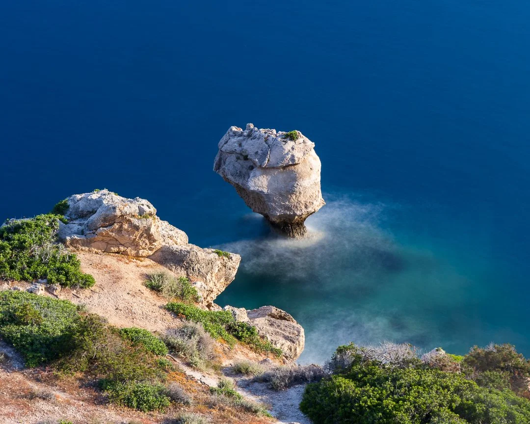 A rocky coastal cliff with green shrubbery overlooking the ocean, featuring a large rock formation rising out of the water.