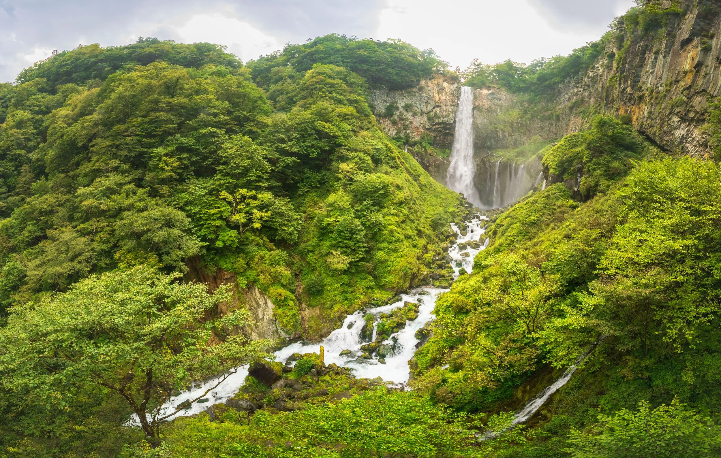 A scenic view of a lush green forested landscape with a waterfall cascading down a cliff, surrounded by trees and flowing stream, under a cloudy sky.