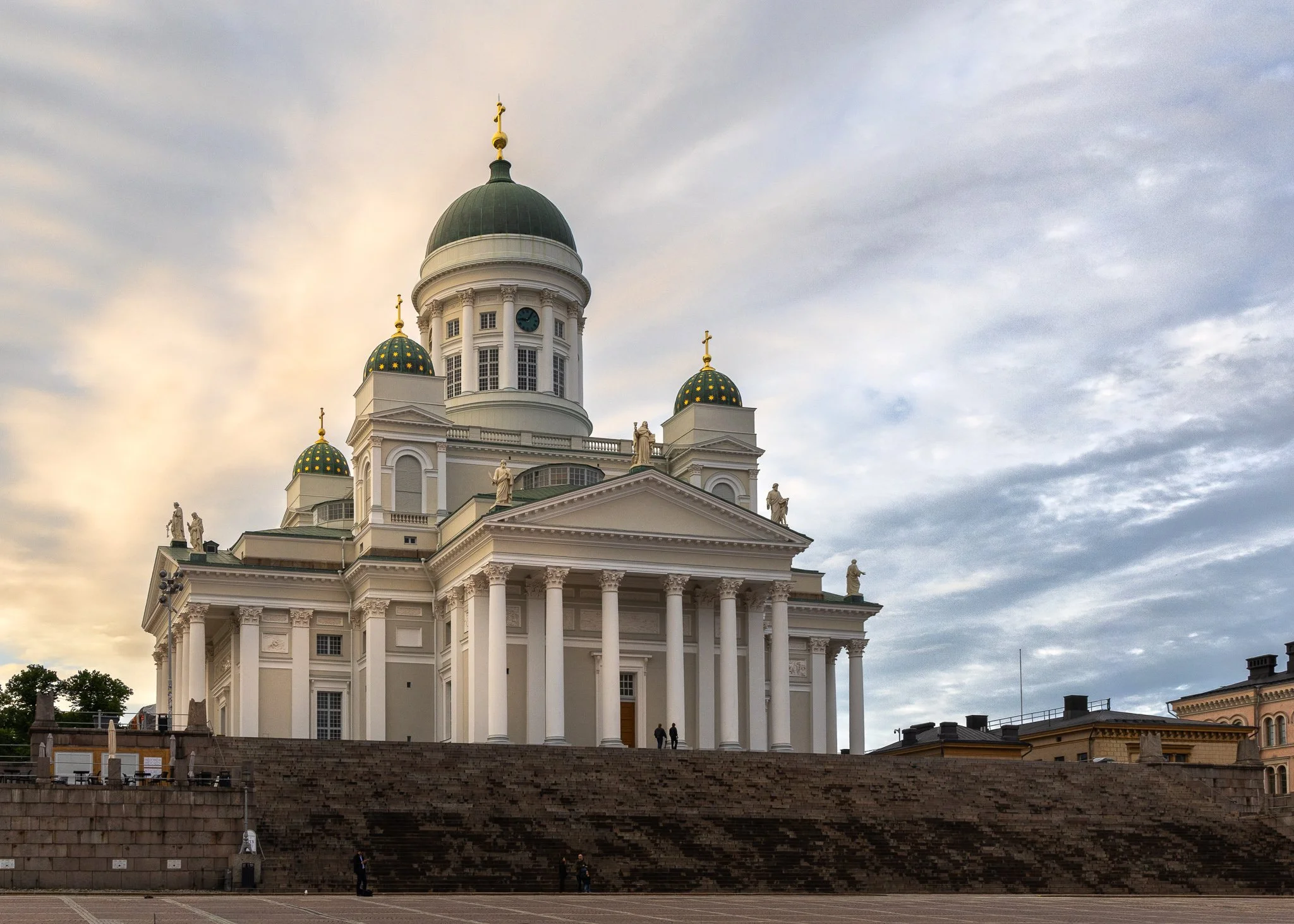 The Helsinki Cathedral, a neoclassical white building with a central green dome and smaller green domes with gold crosses, featuring classical columns and statues on the roof, against a cloudy sky.