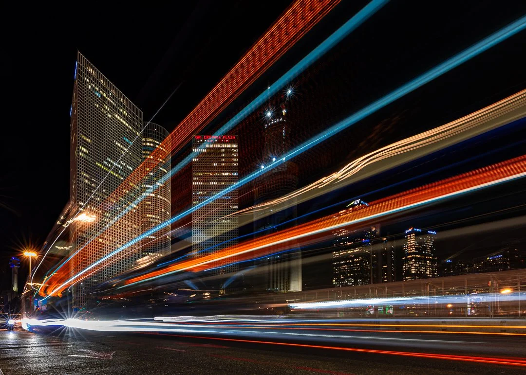 Nighttime cityscape with light trails from moving vehicles and illuminated skyscrapers.