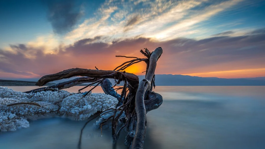 Sunset over a body of water with a large, partially submerged driftwood and salt formations in the foreground.