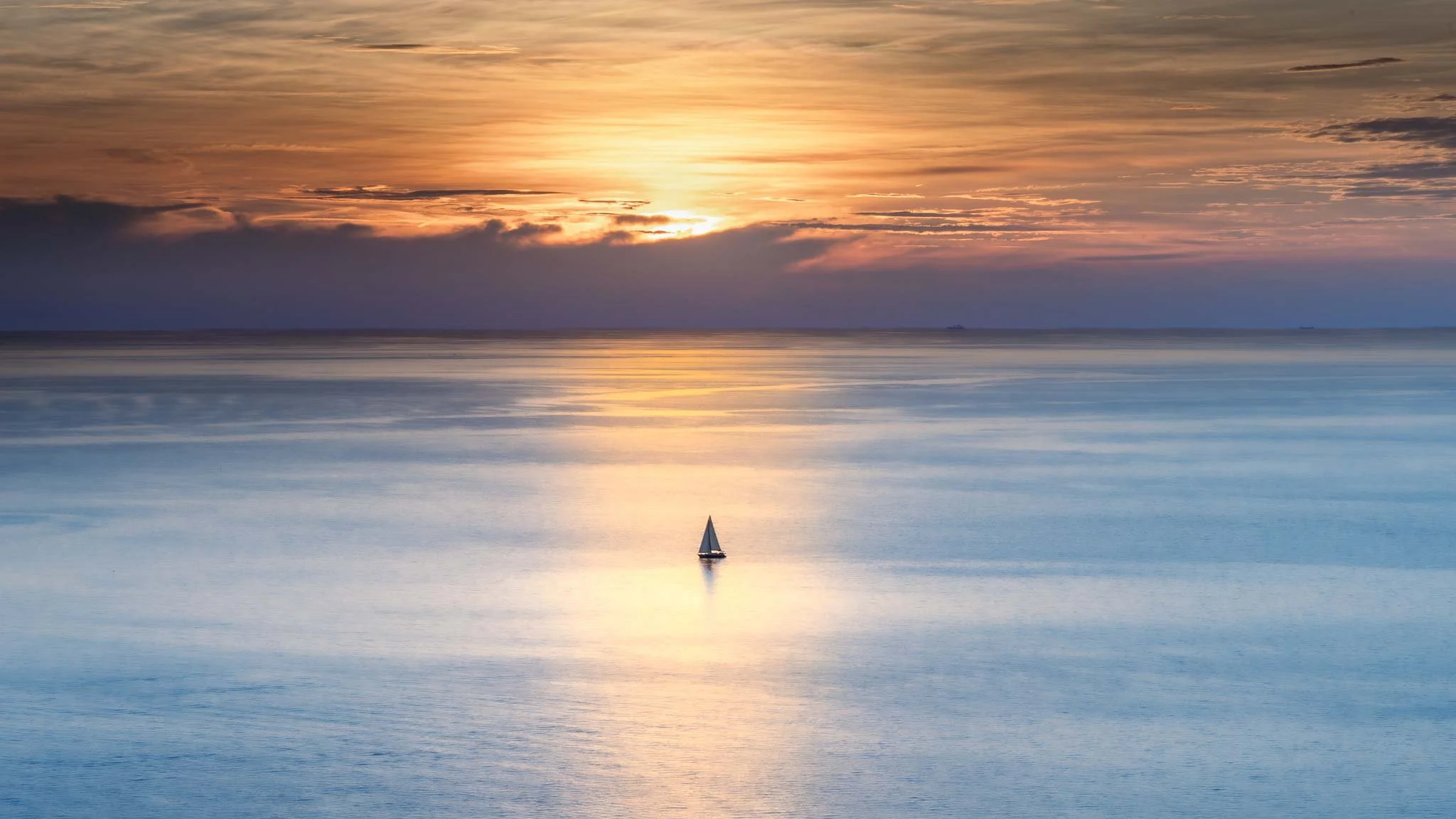 A solitary sailboat crossing a calm blue sea beneath a golden sunset, with warm sunlight reflecting across the smooth water.